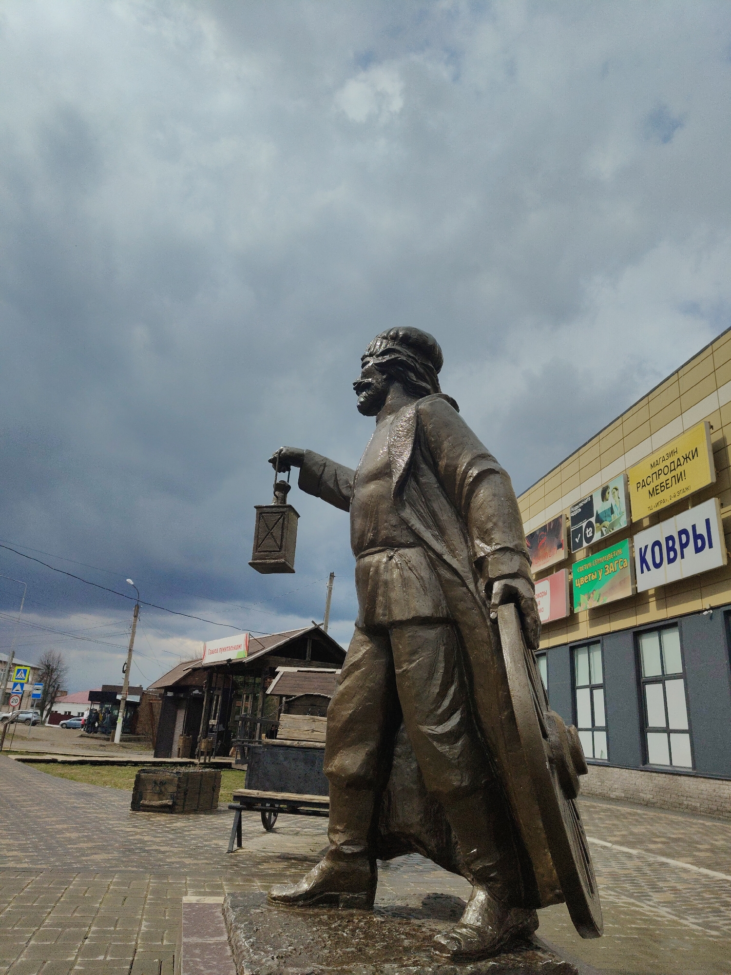 The image features a bronze-colored statue of a man standing prominently outdoors under a heavily clouded, overcast sky. The statue depicts a bearded man wearing what appears to be a long coat or robe and a head covering. He holds an antique-style lantern in his raised left hand and rests his right hand on a large, round object resembling a wheel. The statue is viewed from a low angle, emphasizing its scale against the dramatic sky.
In the background to the right, a modern commercial building stands with light yellow facade panels on its upper section and dark grey walls with white-framed windows below. Numerous advertising signs are attached to its exterior. To the left, a paved sidewalk or road stretches into the distance, with utility poles, power lines, and some older, wooden-clad buildings or stalls visible. A blue road sign with a pedestrian crossing symbol and a "20" speed limit sign are seen on the far left. Near the base of the statue, on the paved ground, are a wooden planter and a metal bench.
The scene combines monumental art with an urban backdrop, featuring a blend of modern commercial architecture and more rustic wooden structures. The dark, cloudy sky suggests an overcast day, lending a somewhat somber or dramatic mood to the setting.
Visible text from the signs includes:
* "МАГАЗИН РАСПРОДАЖИ МЕБЕЛИ!" (Store Furniture Sale!)
* "ТЦ «ИГРА», 2-Й ЭТАЖ!" (Shopping Center "IGRA", 2nd Floor!)
* "КОВРЫ" (Carpets)
* "СВЕТИК-СЕМИЦВЕТИК" (Svetik-Semitsvetik)
* "ЦВЕТЫ У ЗАГСА" (Flowers near the ZAGS)
* "Ганаса пуинтаськом" (on a small red sign)
* "Товары MO te" (below "Ганаса пуинтаськом")
