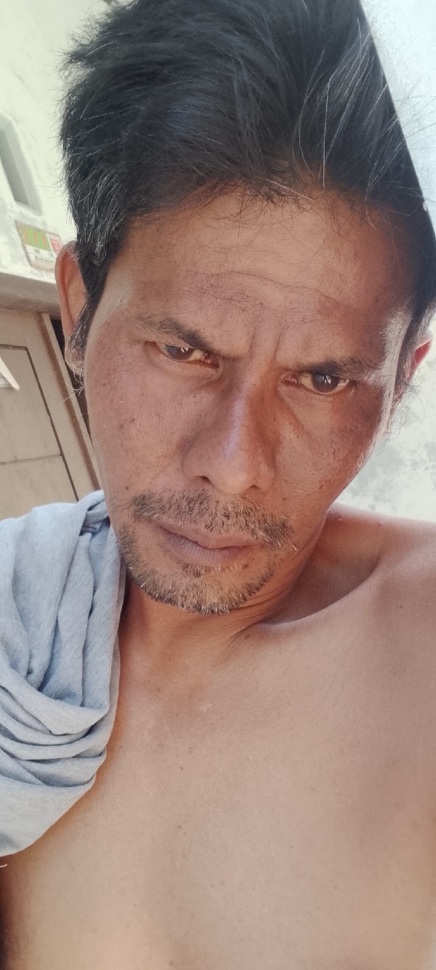 The image is a close-up, selfie-style photograph of an adult male with tanned skin and short, dark hair with some graying. He has a prominent brow, dark eyes that appear to be looking directly at the camera with a serious or intense expression, a dark mustache, and a scruffy beard. The man is shirtless, with a gray t-shirt draped over his left shoulder. The background is blurred, with hints of a building and what appears to be natural light. Based on the contextual information, this photograph was taken in Padang Dalom, Indonesia.