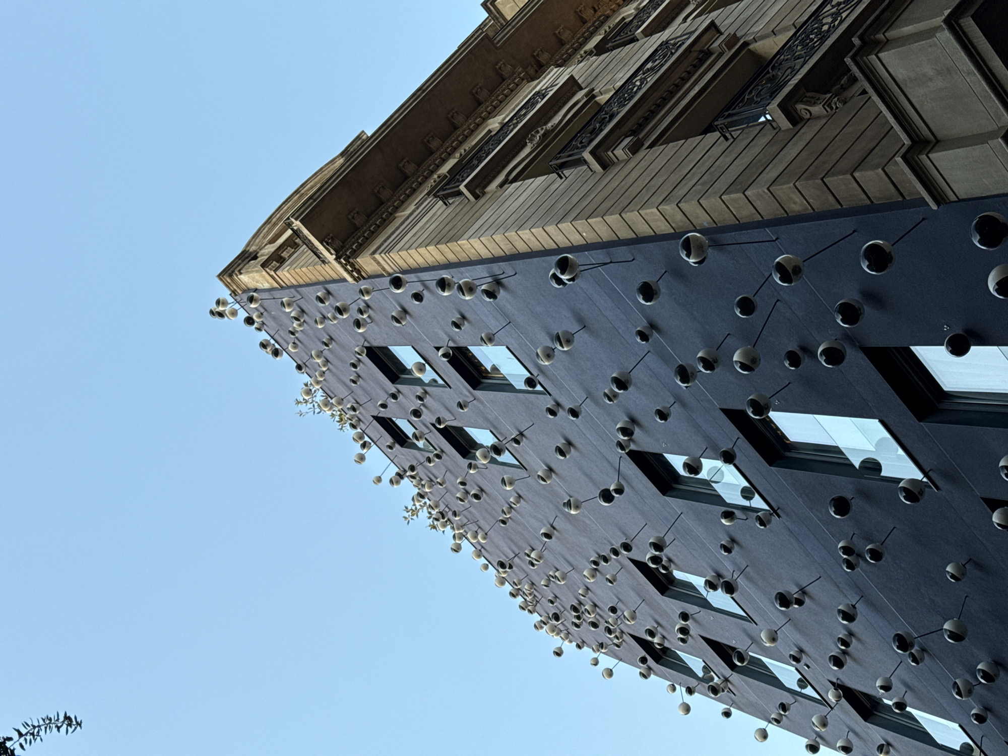 The image is a low-angle shot of a building with a unique facade. The building has a dark-colored exterior with many small, spherical protrusions attached to it by thin rods, creating a textured and somewhat abstract effect. There are also rectangular windows with dark frames evenly distributed across the facade. The building also has a traditionally designed section with balconies and detailed molding at the top. The sky is clear and blue. Based on its unique façade, this building is likely La Casa de les Punxes located in Barcelona, Spain.