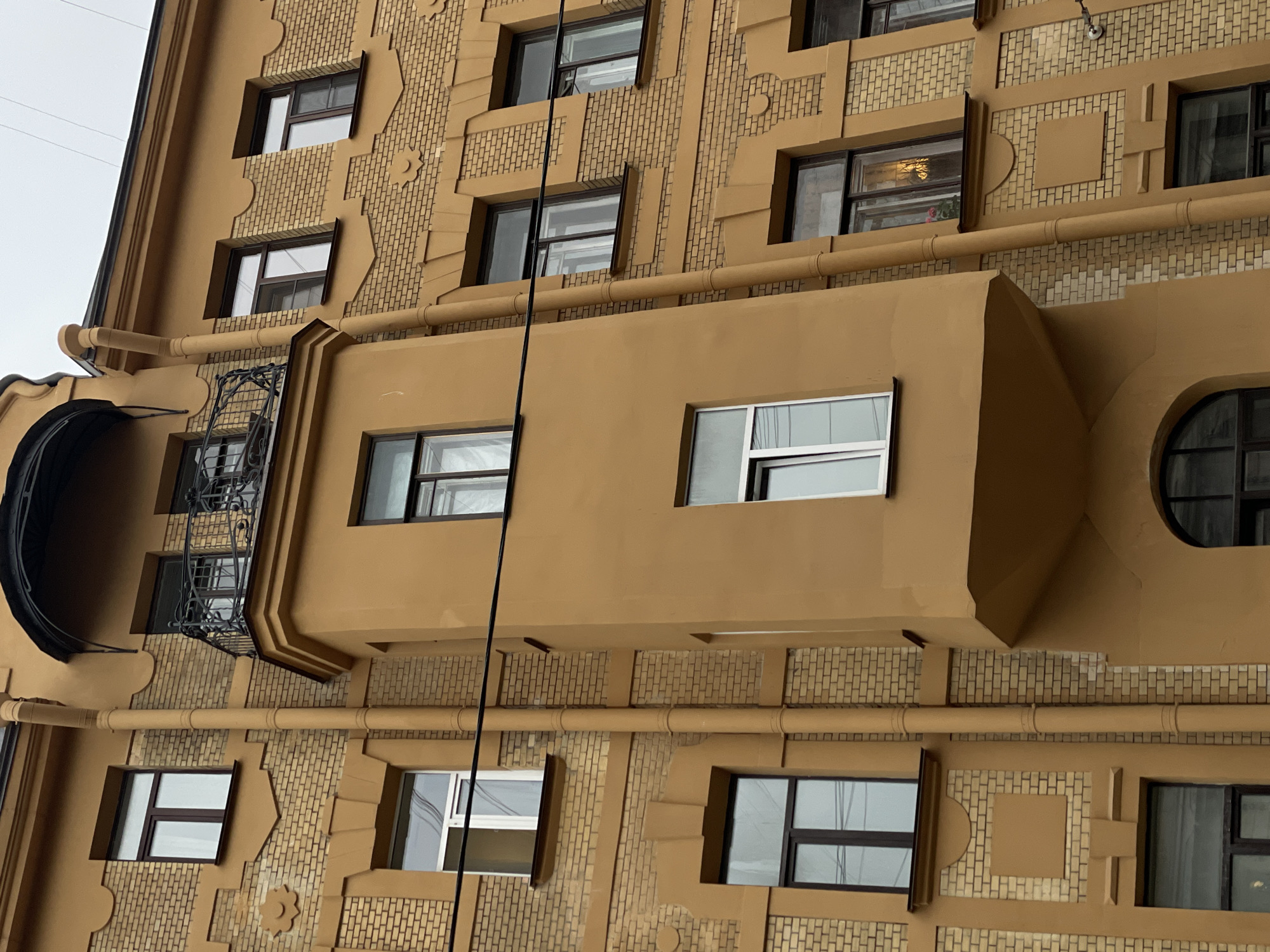 The image shows the exterior facade of a building in Saint Petersburg, Russia. The building is characterized by its yellow brickwork and distinctive architectural style. The facade includes multiple rectangular windows with dark frames, arranged in a somewhat irregular pattern. There are also decorative elements such as protruding ledges and moldings, as well as ornate details around some of the windows. A balcony with black wrought iron railings is visible on the left side of the image. A brown pipe runs horizontally along the building, and a black cable is suspended vertically in front of the facade.