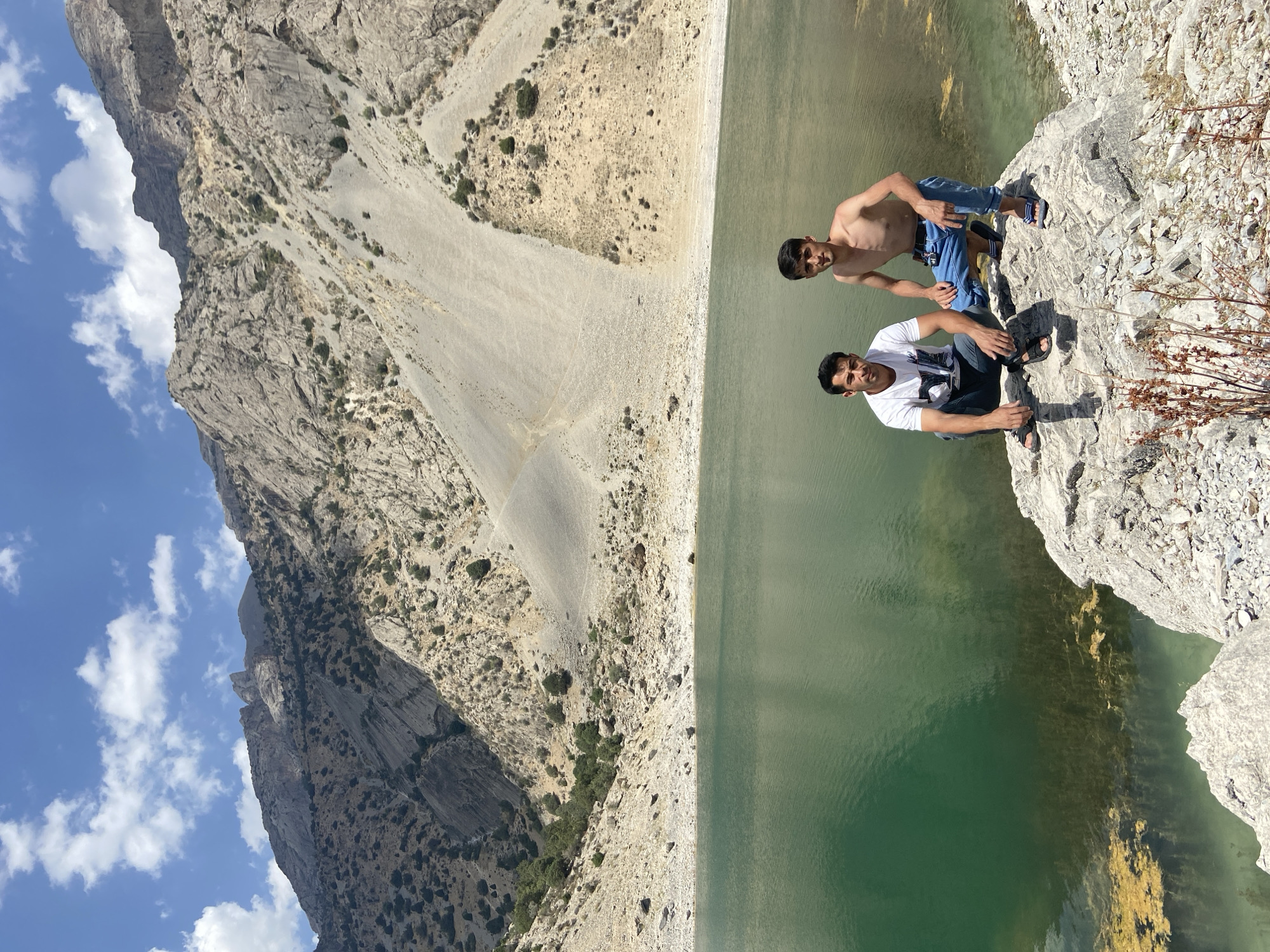 Two young men are posing for a photograph near a calm, green body of water, likely a lake, at the base of a steep, rocky mountain range. The man on the left is wearing a white t-shirt with a dark graphic and blue jeans, while the man on the right is shirtless, wearing blue jeans. Both are crouching on a large, light-colored rock formation that extends into the foreground. The water is clear enough to see submerged vegetation in some areas. The background is dominated by the rugged, barren slopes of the mountains, with patches of sparse vegetation and a bright blue sky with scattered white clouds. The overall scene suggests a remote, natural landscape, possibly in the Pamir Mountains, which are prominent in Tajikistan, including areas near Ghazza.