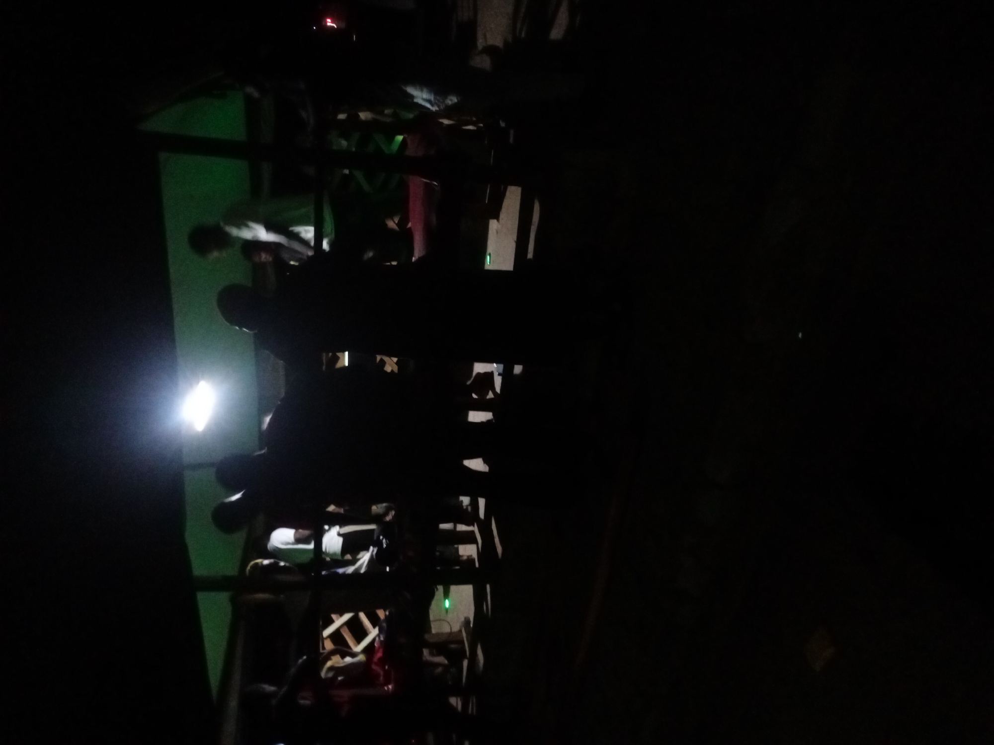The image shows a poorly lit, cluttered outdoor scene at night. A scaffold-like structure is visible, constructed from wood and metal, possibly part of a building under construction or renovation. The structure is filled with various objects, including clothing and other items. A bright light source illuminates a portion of the scene, creating strong contrasts and casting deep shadows. The location is Unknown, Unknown. No specific details about people, actions, or interactions are discernible due to the poor lighting and obstructed view.