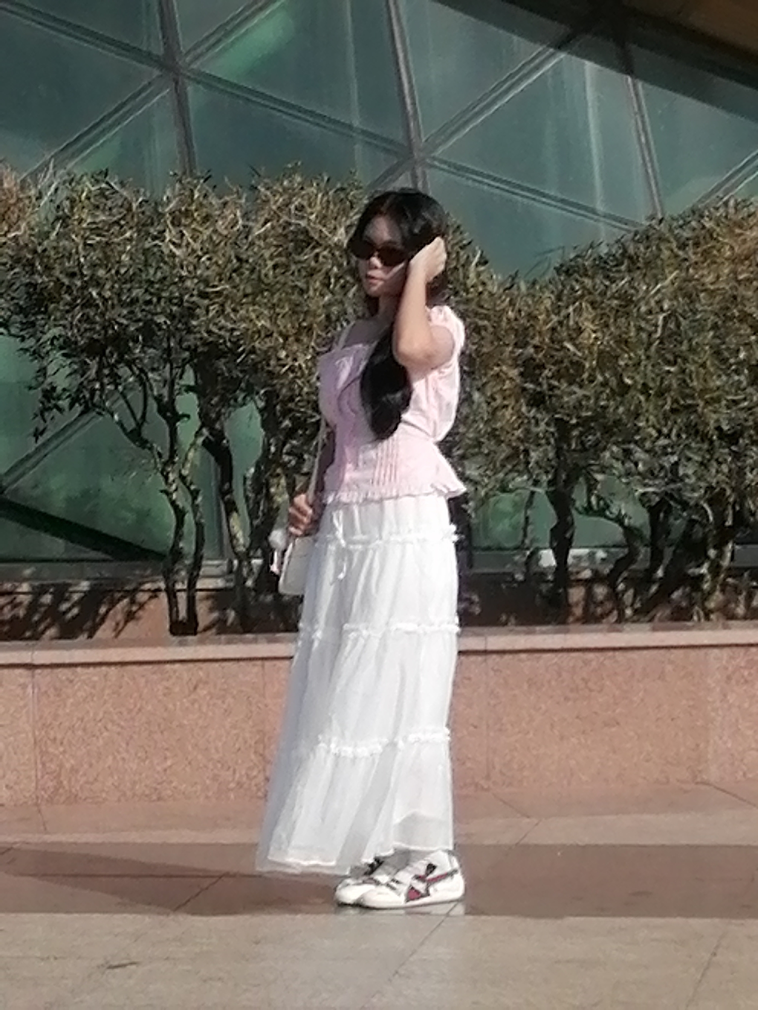 A young woman stands in a full-length, tiered white skirt and a pink off-the-shoulder top. She wears sunglasses and has her hand on her head. She carries a white purse on her left shoulder. She is wearing white sneakers with red and black accents. The background features lush green trees and a textured brown wall. The setting appears to be an outdoor plaza in Da Lat, Vietnam, with a distinctive geodesic dome-like structure visible in the background.