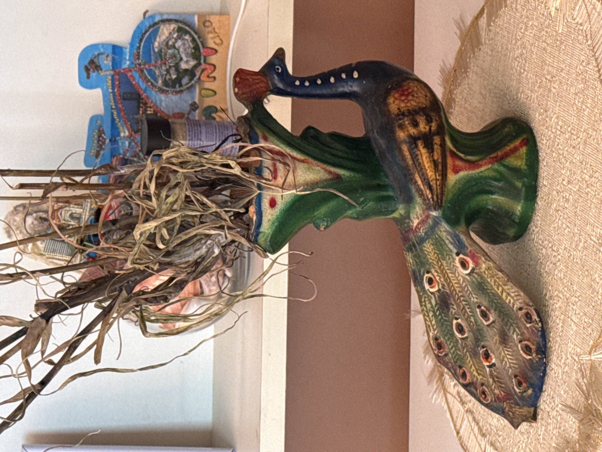 A ceramic peacock figurine, painted in various colors including green, blue, yellow, and red, stands on a textured placemat. The peacock holds dried plant stalks in its back, which sprout from a green, vase-like structure. The figurine is placed on a white surface, with a brown wall visible in the background. Behind the figurine, a colorful cardboard cutout of a bicycle with mountains and the word "CIAO" is visible. A small, dark-lidded bottle is also partially visible to the left of the cutout. The scene appears to be an indoor setting, possibly a shelf or tabletop, but the specific location in Montana, Bulgaria, cannot be determined from the image.