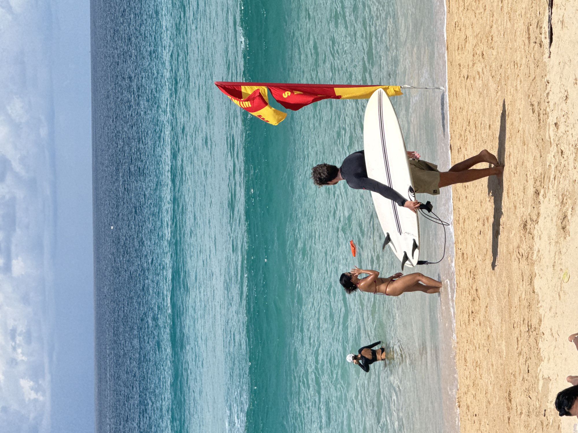 The image shows a beach scene with the ocean in the background. In the foreground, a person wearing a black wetsuit and shorts is walking on the sand, carrying a white surfboard. To the right of this person, a red and yellow flag is planted in the sand. In the shallow water, two people are present. One is wearing a black wetsuit and a white hat, and the other is wearing a bikini. A red float is visible in the water near the person in the bikini. The water is a clear turquoise color, and the sky is overcast with clouds. The sand is light brown and appears wet near the water's edge. The overall impression is of a bright, sunny day at the beach, possibly with moderate surf.