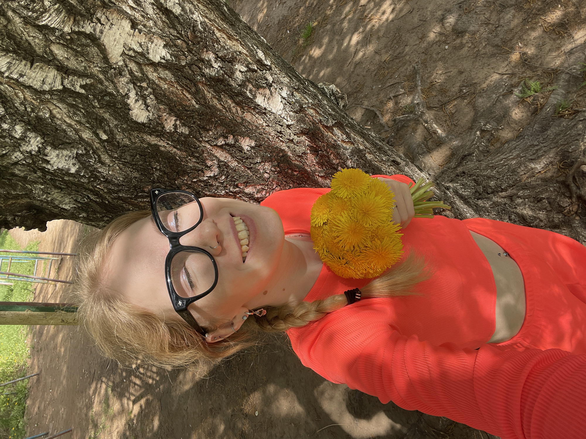 A fair-skinned woman lies on her back against a tree in Izhevsk, Russia. She wears large black-rimmed glasses and a coral-colored ribbed long-sleeved top with a cutout on the left side revealing her abdomen and a belly button piercing. She holds a bouquet of dandelions near her left shoulder. Her blonde hair is partially braided, with the braid lying across her shoulder. The tree trunk is thick and textured. The ground is covered with dirt and tree roots. A playground structure is visible in the background on the left side of the frame.