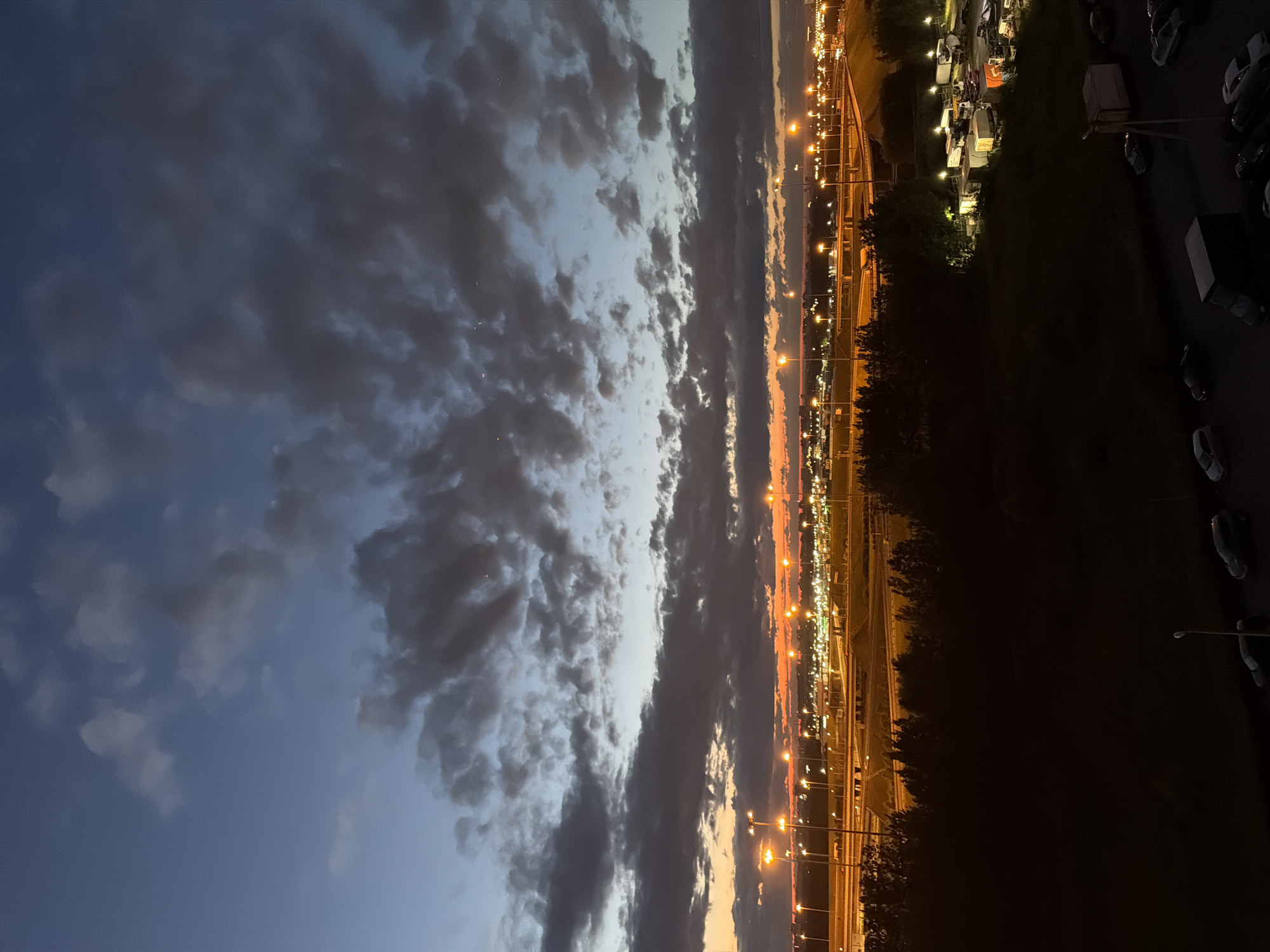 An aerial perspective captures a twilight scene over a city landscape. The sky is a gradient of deep blue and dark grey clouds, with lighter shades of orange and yellow on the horizon, indicating the last light of day. Streaks of orange light emanate from numerous street lamps illuminating a complex network of roads and overpasses in the mid-ground. The roads are lined with trees on either side. In the foreground, a dark, silhouetted area of trees and foliage separates the road system from a parking lot filled with various vehicles, including trucks, vans, and cars. The parking lot is dimly lit by scattered lights, and some of the vehicles appear to be parked trailers. The overall scene suggests an urban or industrial area at dusk.