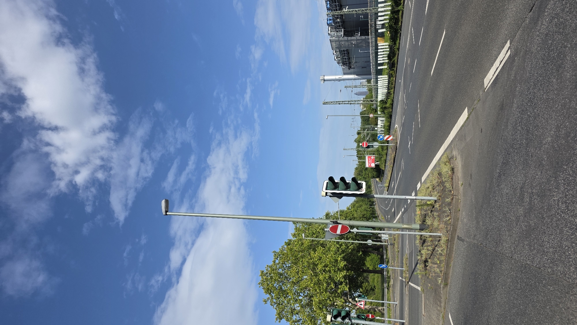 The image shows a street scene in an urban environment. The foreground is dominated by an asphalt road with white lane markings. On the left side of the road, there is a traffic light with a red signal illuminated. Further back, another traffic light is visible, with all three lights (red, yellow, and green) illuminated. Several traffic signs are also present, including a round red and white prohibitory sign and a blue circular sign with a white arrow. A tall street lamp stands prominently on the left side of the image. In the background, a large green tree is visible, and beyond that, a complex industrial building with a tall smokestack. The sky is blue with scattered white clouds. The overall impression is that of an active intersection on a clear day.