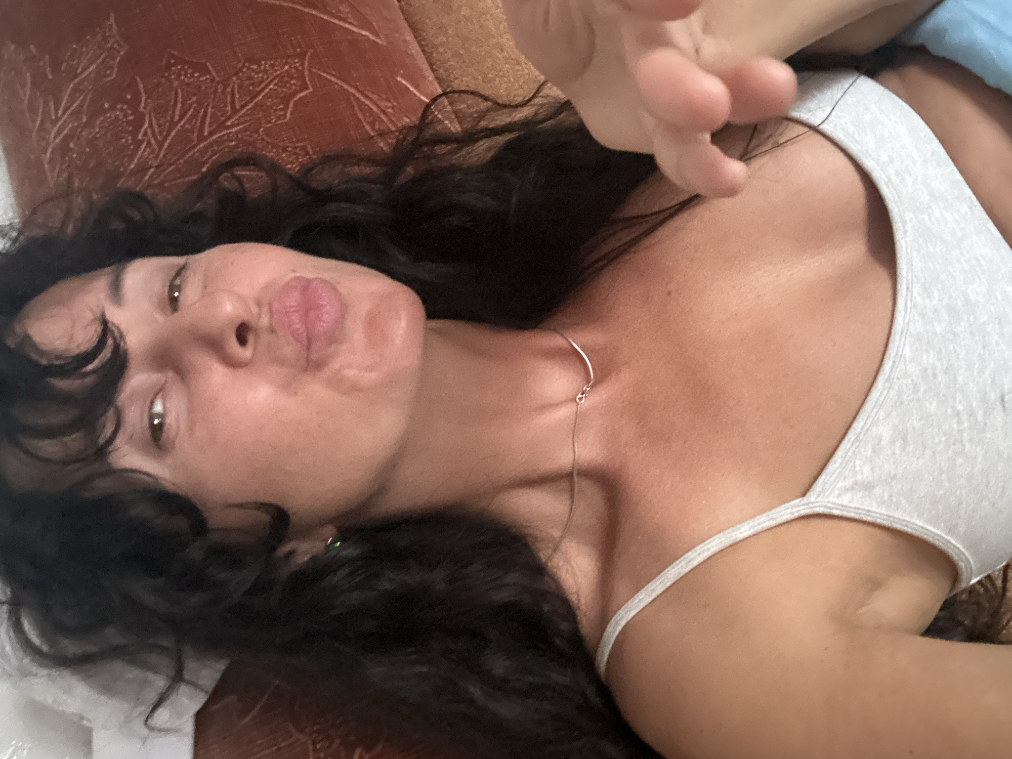 A woman with dark, curly hair and bangs is lying down, looking at the camera with a playful expression, puckering her lips. She is wearing a light gray tank top and a thin silver necklace. Her right hand is raised towards the camera, fingers slightly curled. She has a green earring in her left ear. The background appears to be a textured, reddish-brown surface, possibly a couch or pillow. The lighting is soft, and the image appears to be taken indoors. There is no discernible information about the location in Shelekhov, Russia, within the image itself.