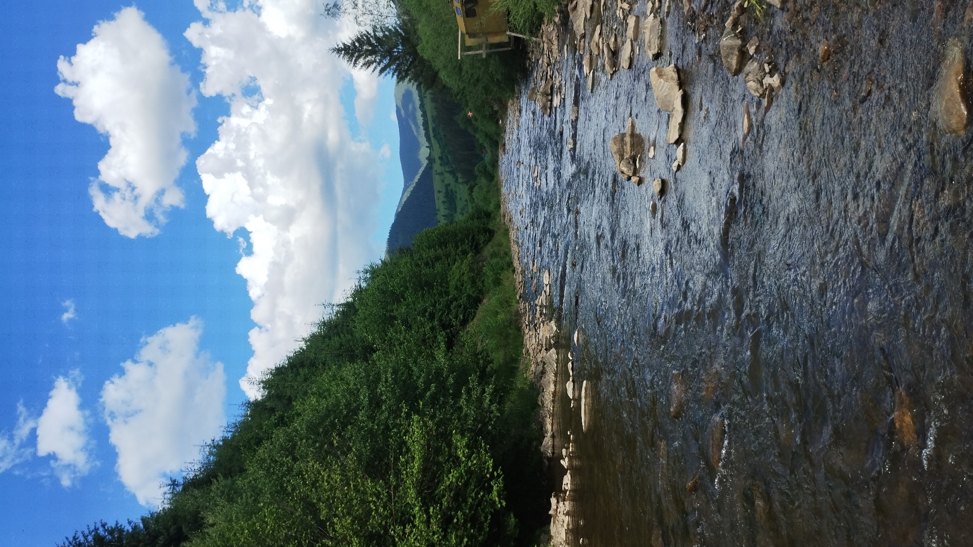 The image depicts a scenic view of a river flowing through a mountainous region. The river, with its rocky bed and clear water, is the central focus. Lush green trees line the left bank, forming a dense forest that extends up the hillside. On the right bank, a yellow structure, possibly a small building or cabin, is visible amongst the trees. In the background, rolling hills covered in vegetation and distant mountains under a bright blue sky with scattered white clouds complete the landscape. The scene suggests a natural, undeveloped area, typical of a rural or wilderness setting.