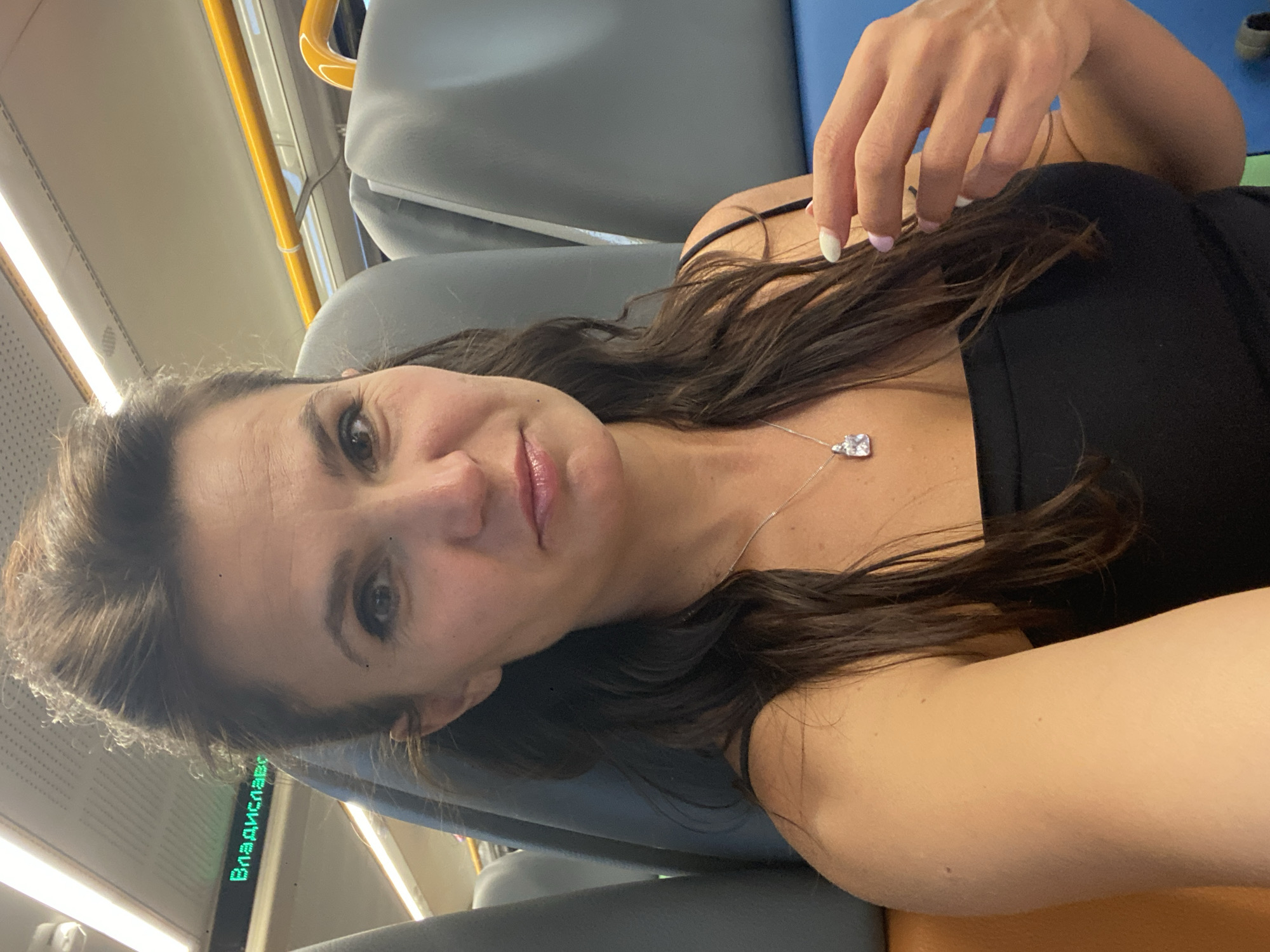 A woman with brown hair is taking a selfie on a train. She is wearing a black tank top and a silver necklace with a diamond pendant. Her left hand is touching her hair. The train has gray seats and yellow handrails. A digital display above her head shows the word "Владислав" in green letters, indicating the train's destination is Vladyslavovske rural settlement in Ukraine. The lighting suggests it is daytime.