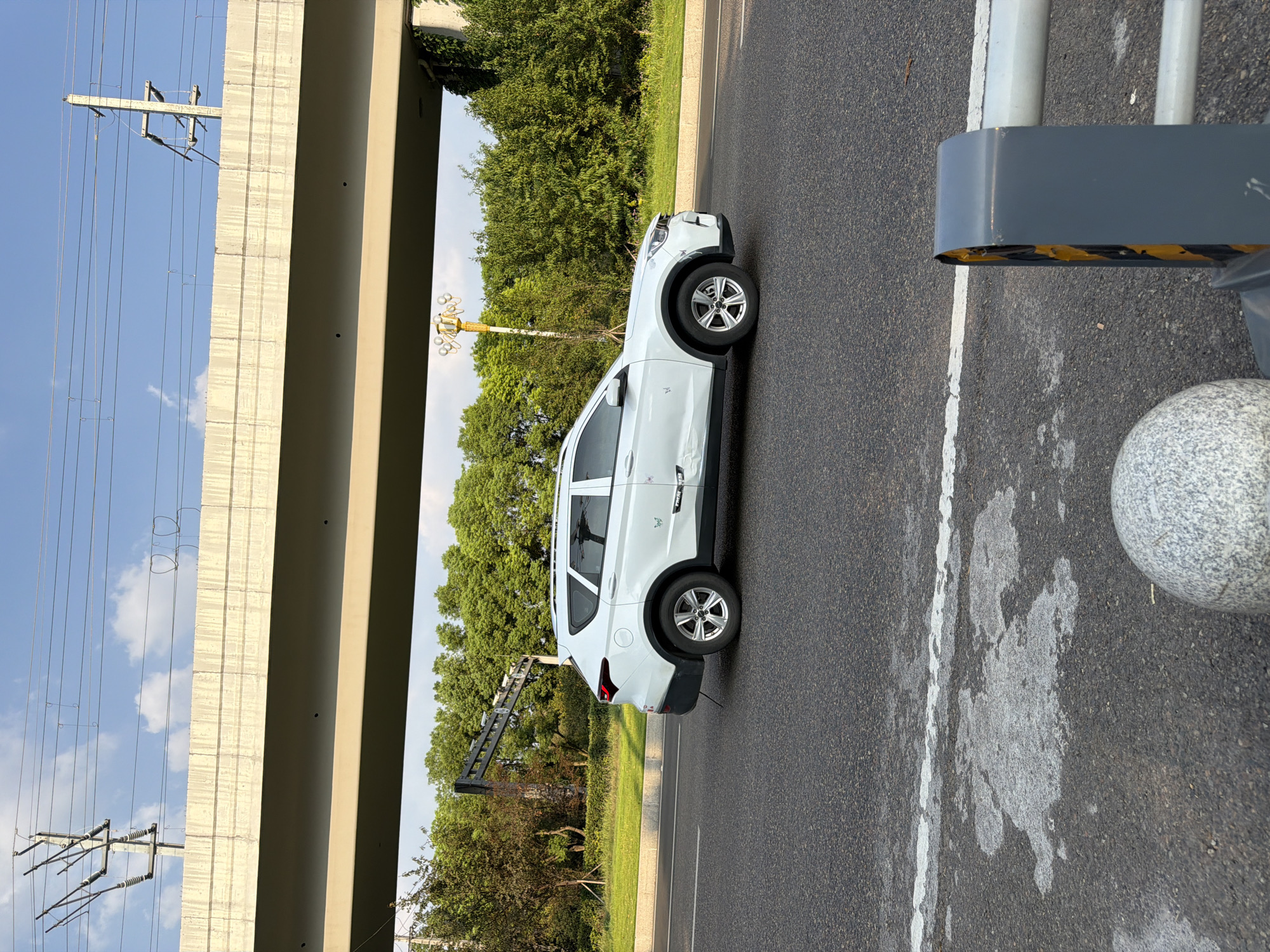 In Jiangjin, China, a white SUV is traveling on a paved road under a concrete overpass. The SUV has damage to its side, including a dent and what appears to be a scratch. The road has white lane markings, and the surrounding area is lined with green trees and grass. Overhead, power lines and a utility pole are visible against a blue sky with some clouds. In the foreground, a gray, textured sphere and a dark gray barrier with yellow accents are present.