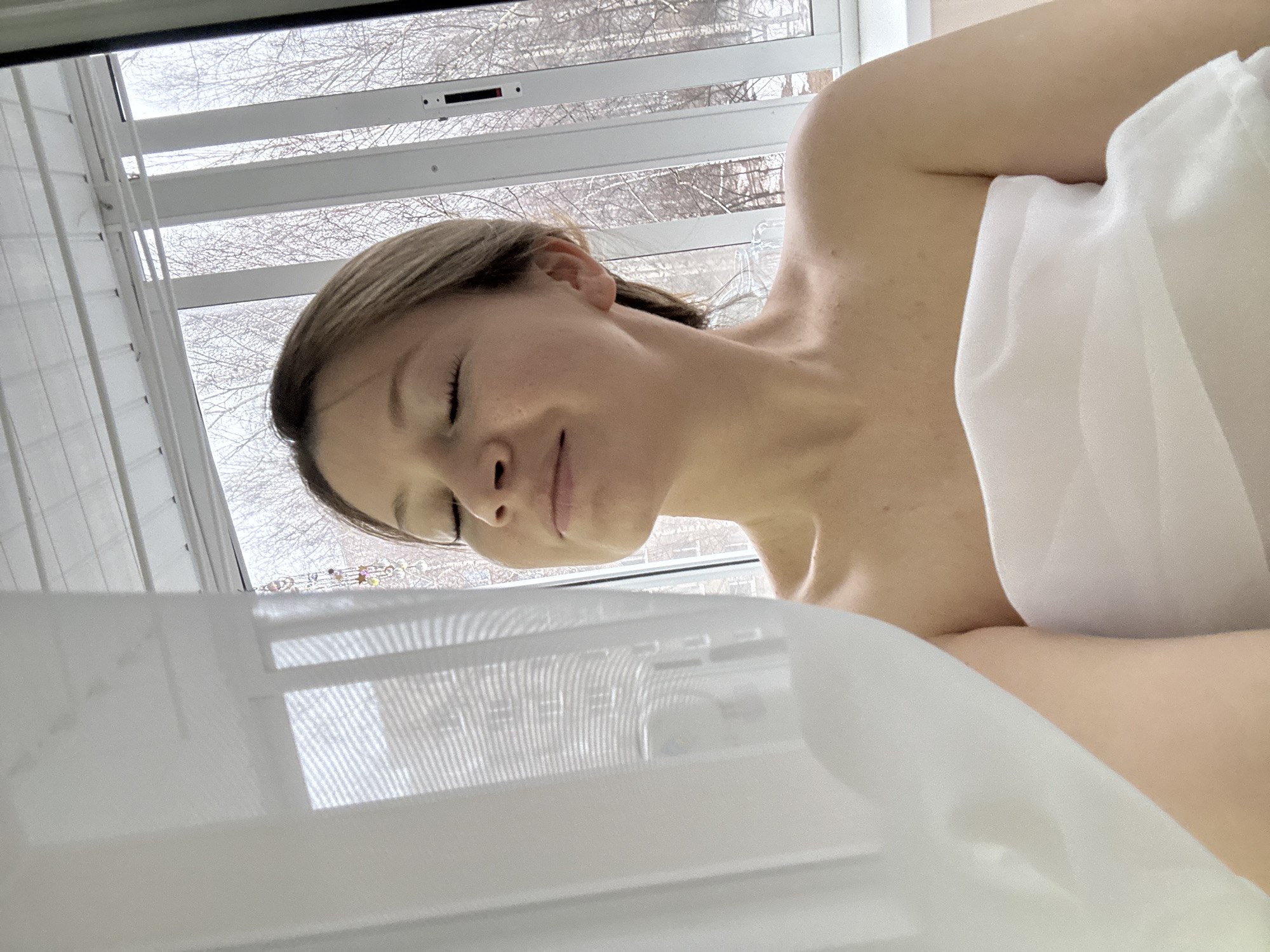 A woman is lying down, wrapped in a white sheet. She has fair skin, short light brown hair, and her eyes are closed. She appears to be indoors, near a window with white blinds. Through the blinds, bare trees are visible. The location is likely Chaikovsky, Russia, based on implied context. The scene suggests a peaceful, intimate moment of relaxation or rest.