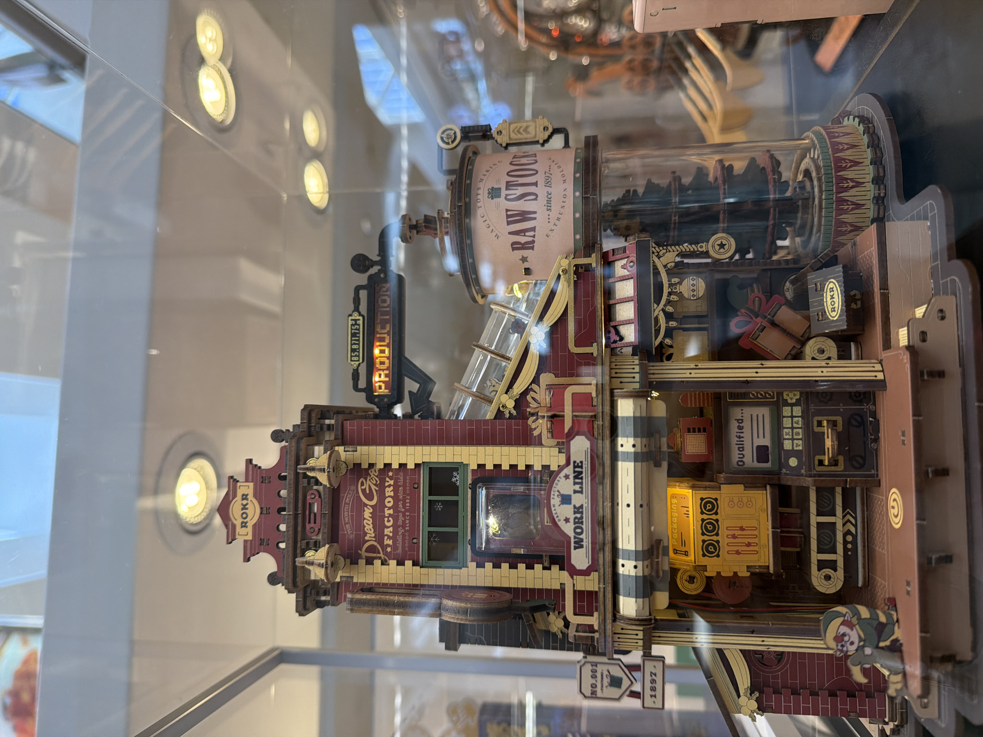 The image shows a detailed model of a factory, possibly a toy factory, displayed behind glass. The factory is multi-storied and made of wood, with intricate details and signage. Prominent labels include "ROKR," "Dream Glow FACTORY," "PRODUCTION," "RAW STOCK," and "WORK LINE." There are miniature figures, machinery, conveyor belts, and decorative elements like gift boxes and a Christmas tree inside a glass dome. The overall aesthetic suggests a whimsical and vintage theme. The model appears to be part of a retail display, as indicated by the reflections of overhead lighting and display cases in the background. The location is Mission Viejo, United States of America.