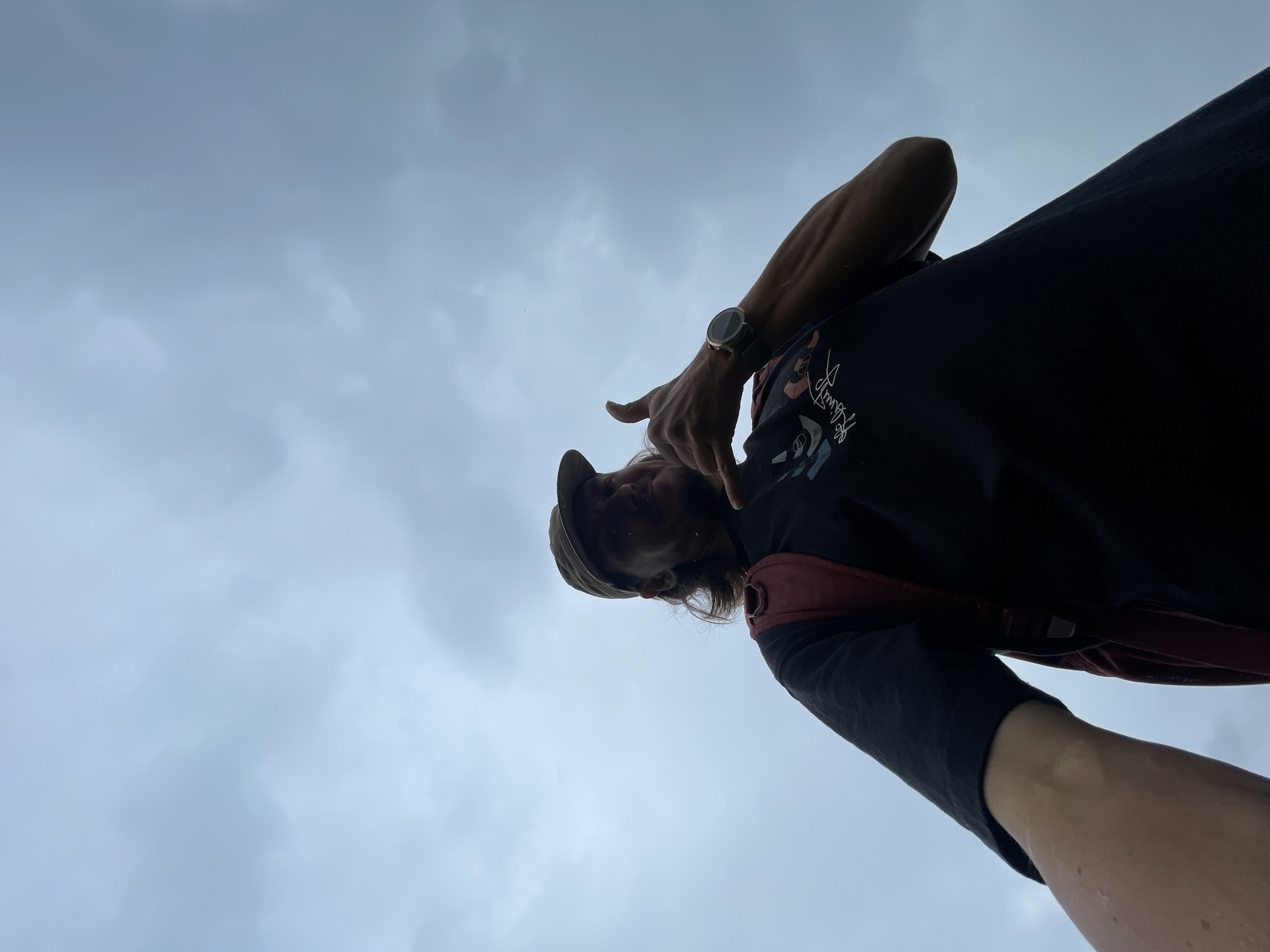 A low-angle, wide shot captures a person from the chest up, silhouetted against a cloudy sky. The individual is wearing a black t-shirt with a graphic print and a red backpack. A baseball cap is worn backward on their head, and their hair is visible hanging down. Their right arm is extended, and their hand is in a "shaka" gesture, with a watch on their wrist. The background is dominated by a light gray, overcast sky with subtle cloud formations.