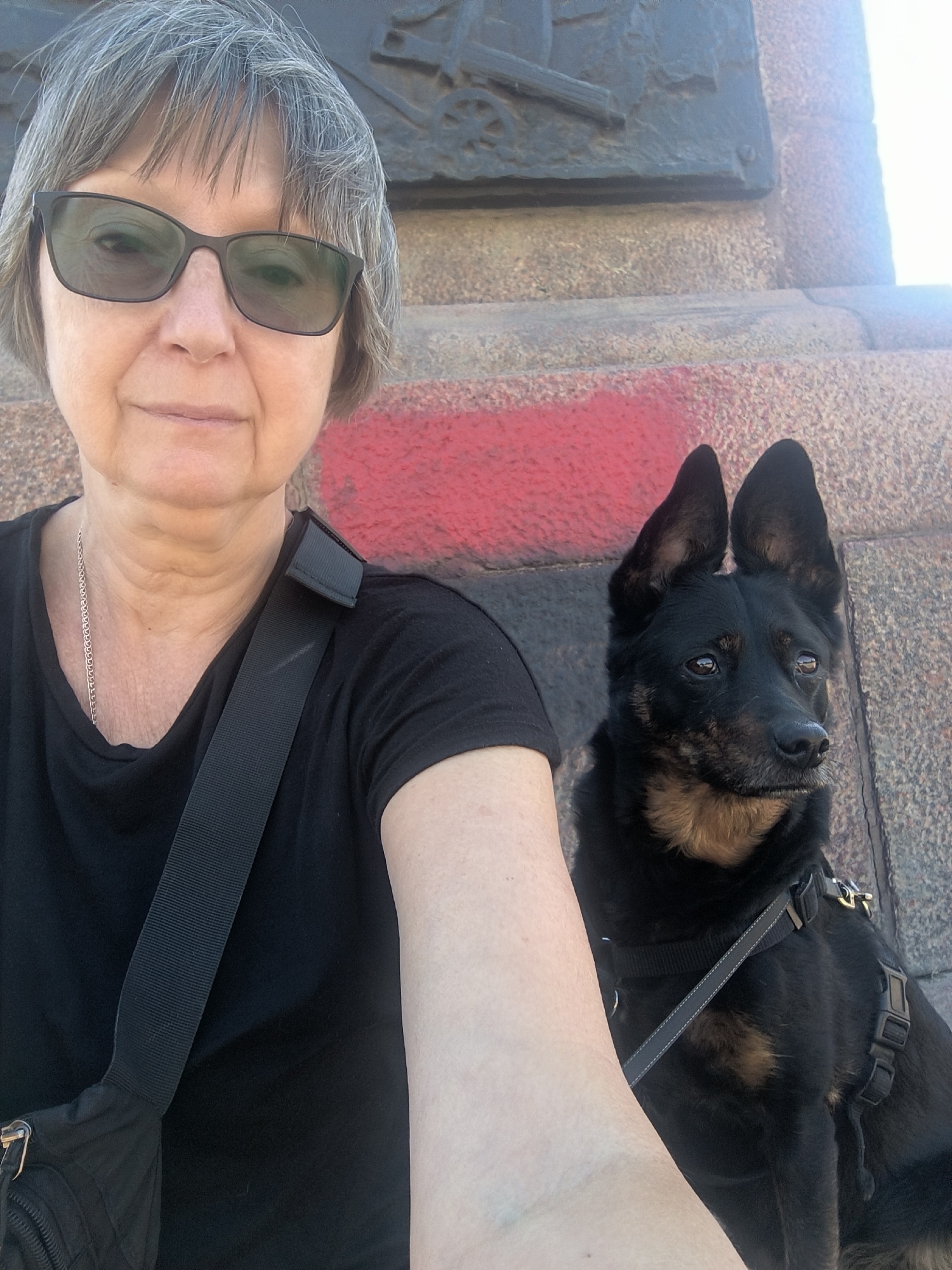 A selfie shows a woman and a black dog outdoors. The woman, wearing sunglasses and a black t-shirt, is on the left side of the frame, with her arm extended. The dog, a medium-sized black dog with tan markings and pointed ears, is seated on the right. They are positioned in front of a stone structure with a red painted section. The background appears to be a textured stone monument or building. The lighting suggests it is daytime, with some glare visible. The context provided about the location in Mykolaiv, Ukraine, is not visually discernible from the image itself.