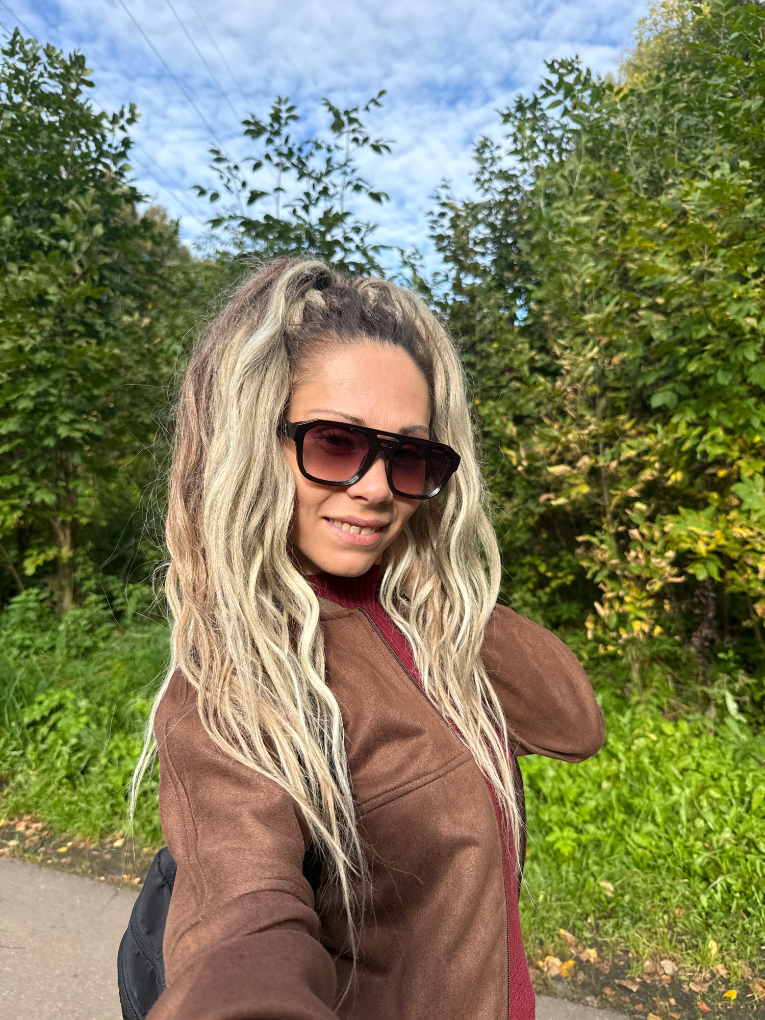 In Rusanovo, Russia, a woman with long, blonde dreadlocks and stylish sunglasses poses for a selfie outdoors. She is wearing a brown jacket over a dark red turtleneck. The background is filled with lush green trees and a bright blue sky with scattered clouds. A paved path is visible to her left, and some fallen leaves suggest it is autumn. Power lines are visible in the upper part of the image.
