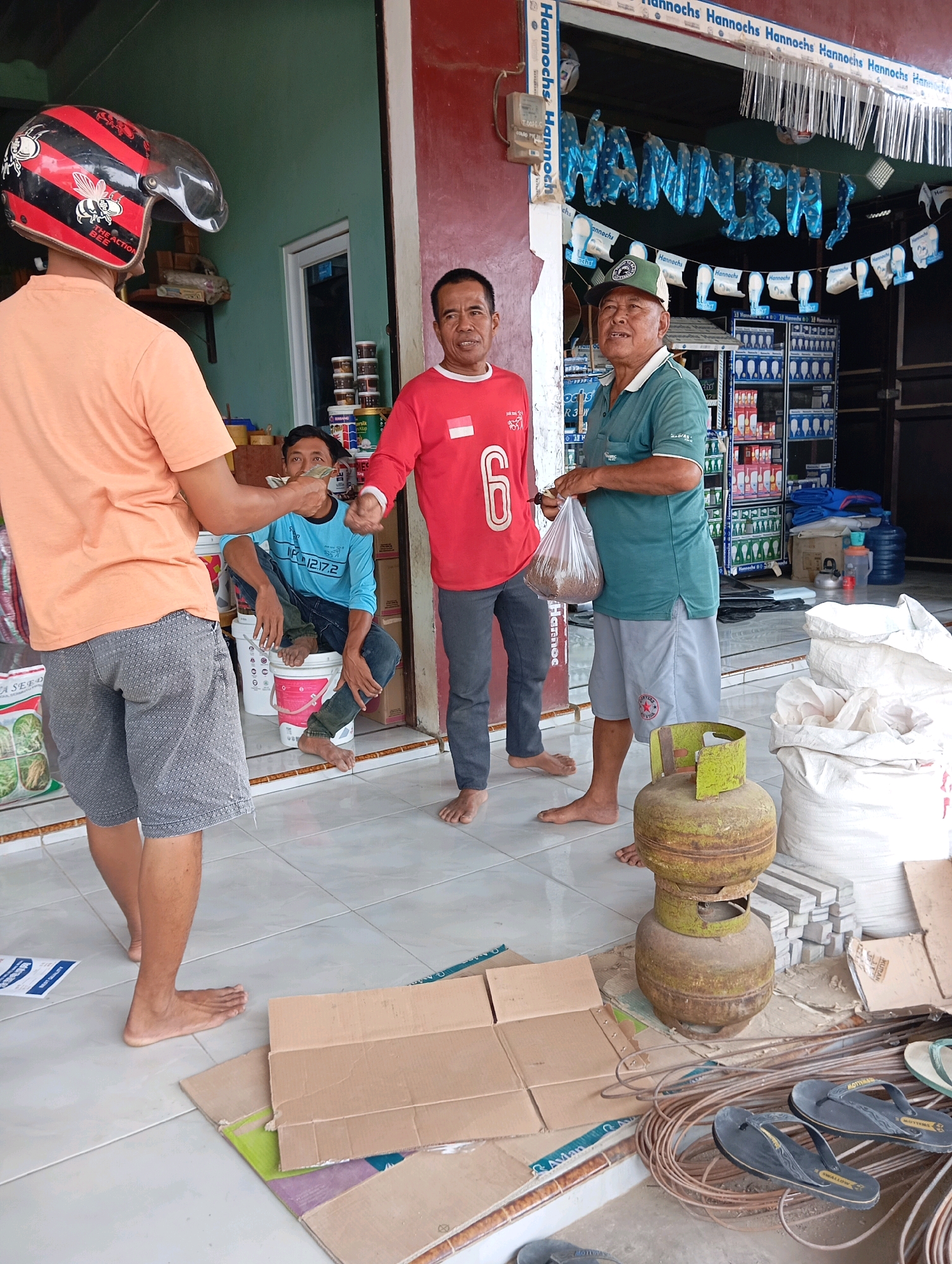 The image captures a scene outside a storefront in Leman Benawi, Indonesia. In the foreground, a person wearing an orange t-shirt and patterned grey shorts stands with their back to the camera, facing away from the viewer. To their left, a man in a red shirt and grey pants is engaged in an exchange with another man sitting on a bucket. This seated man, wearing a blue long-sleeved shirt and dark pants, is holding money. To the right of the seated man, another man in a green polo shirt and grey shorts is holding a plastic bag filled with a granular substance. In front of this man, a rusty green gas cylinder rests on the tiled floor, surrounded by various items including coiled wires, stacks of what appear to be concrete slabs, and large white sacks. The storefront itself has a sign with the name "Hannochs" displayed prominently across it, and shelves stocked with products are visible inside. The overall impression is one of a transaction taking place in a commercial area.