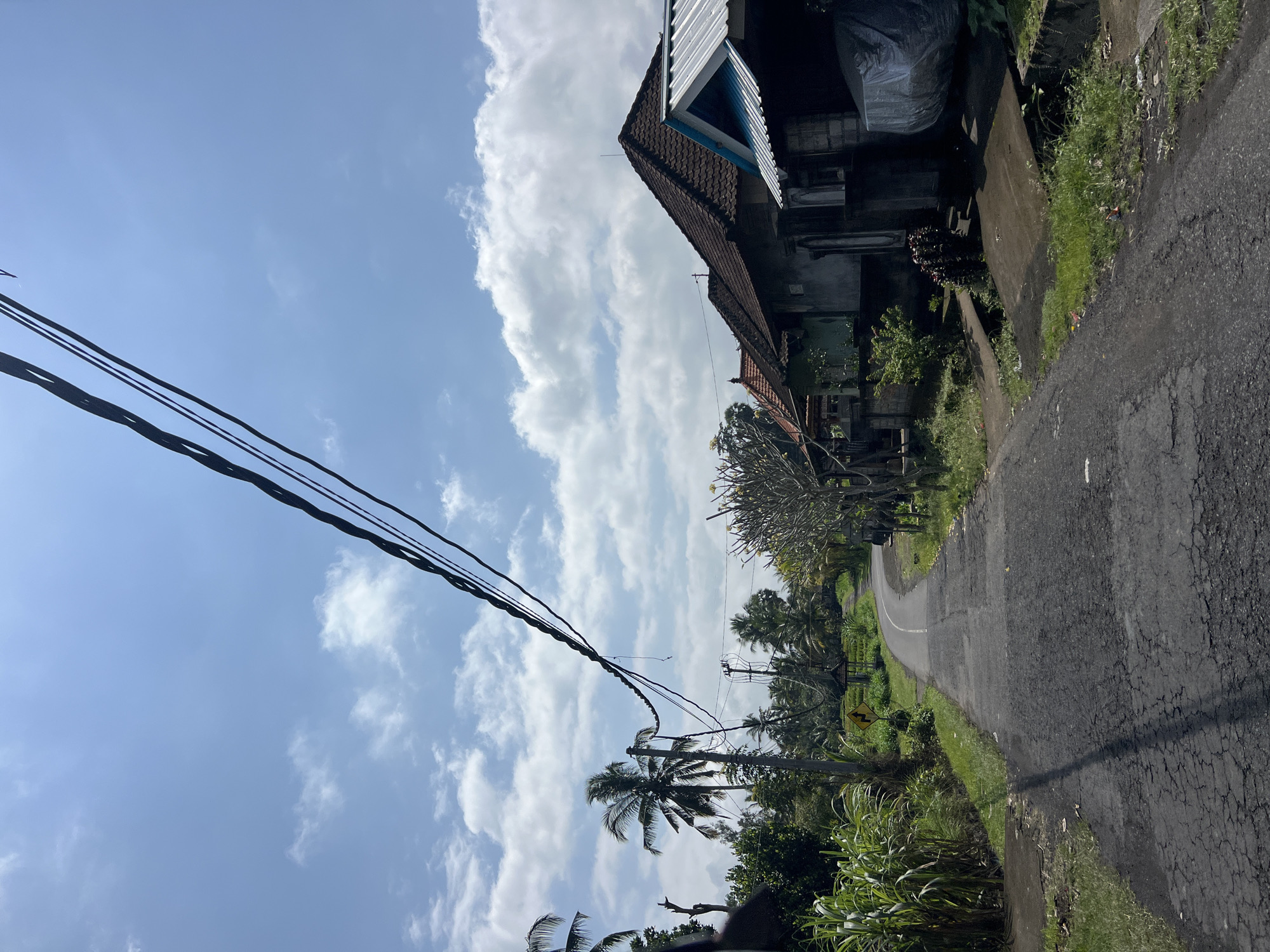 The image depicts a rural road in Susut, Indonesia, on a clear day with a bright blue sky and scattered white clouds. The road is paved and cracked, winding through a lush green landscape. On the right side of the road, several houses with traditional tiled roofs are visible, some with modern corrugated metal roofing. In front of one house, a vehicle is covered with a plastic tarp. Tall green plants, possibly sugar cane, line the left side of the road. Palm trees and other tropical vegetation are abundant throughout the scene. Electrical wires hang prominently across the sky, supported by wooden poles. A yellow sign with black arrows indicating a curve in the road is posted on the left side. The overall impression is one of a peaceful, tropical rural setting.