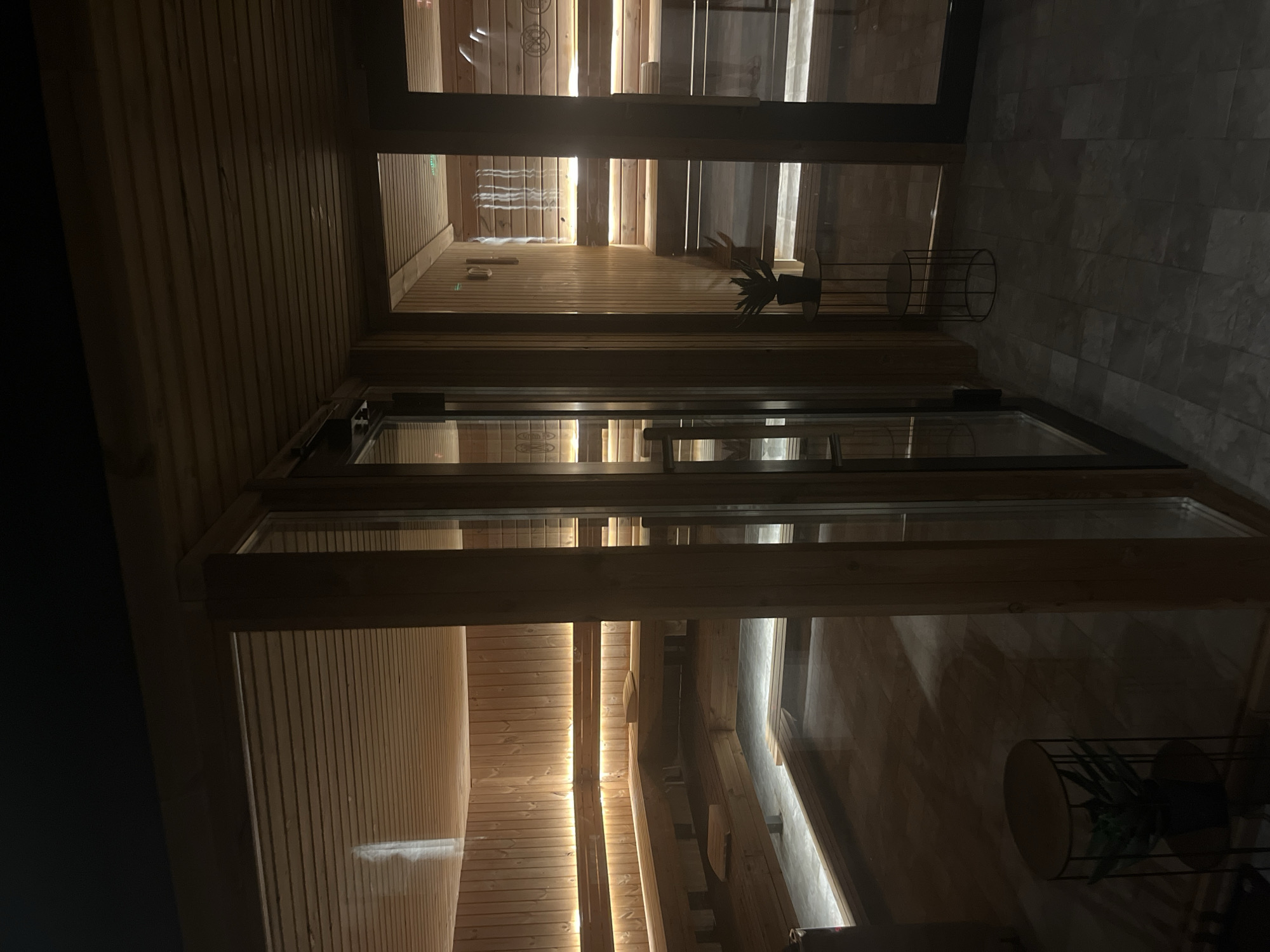 The image captures the interior of a room with wood paneling and integrated lighting. The room has what appears to be a glass-enclosed sauna or similar relaxation space. The lighting emphasizes the wooden slats and creates a warm ambiance. The view is angled, showing the ceiling and an adjacent gray tiled wall. A plant is visible on a stand. The location is unspecified, but the mention of Sofia, Bulgaria, suggests the photo might have been taken there. The details focus on the architectural elements and the lighting design. No people are visible.