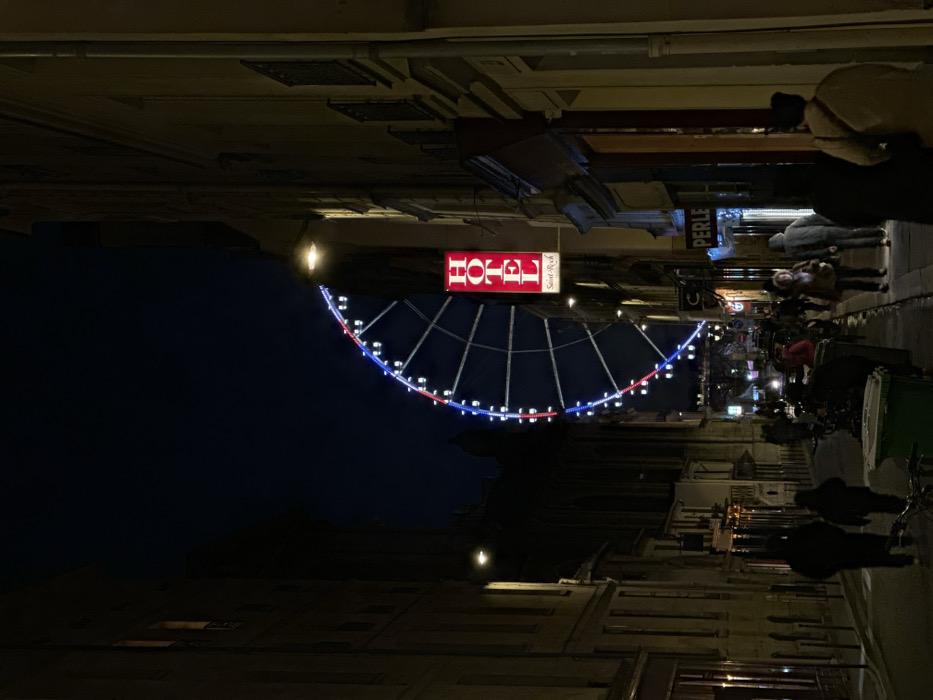 The image depicts a narrow street in Paris, France, at night, viewed from a slightly elevated perspective looking down the thoroughfare. The scene is dimly lit by various artificial sources, casting long shadows and highlighting architectural details.
A striking red, rectangular sign with "HOTEL" written vertically in white letters, and "Saint-Roch" beneath it, is prominently displayed on the right side of the street. Below this, another sign with "PERLE" is partially visible. Stretching across the street, connecting the buildings, is a large, semi-circular arch of decorative lights featuring red, white, and blue bulbs, adding a festive glow to the dark environment.
The street is lined with multi-story buildings typical of Parisian architecture, their windows and facades illuminated in patches. Several people are visible on the street, mostly in the mid-ground and background, appearing as dark silhouettes or blurred figures due to the low light and potential movement. Some individuals are standing or walking on the right side of the street. Street furniture, including a green bin and what appear to be parked bicycles or scooters, are also visible along the right curb. The time is clearly night, with the sky a deep black. The overall atmosphere suggests a quiet yet active urban evening.