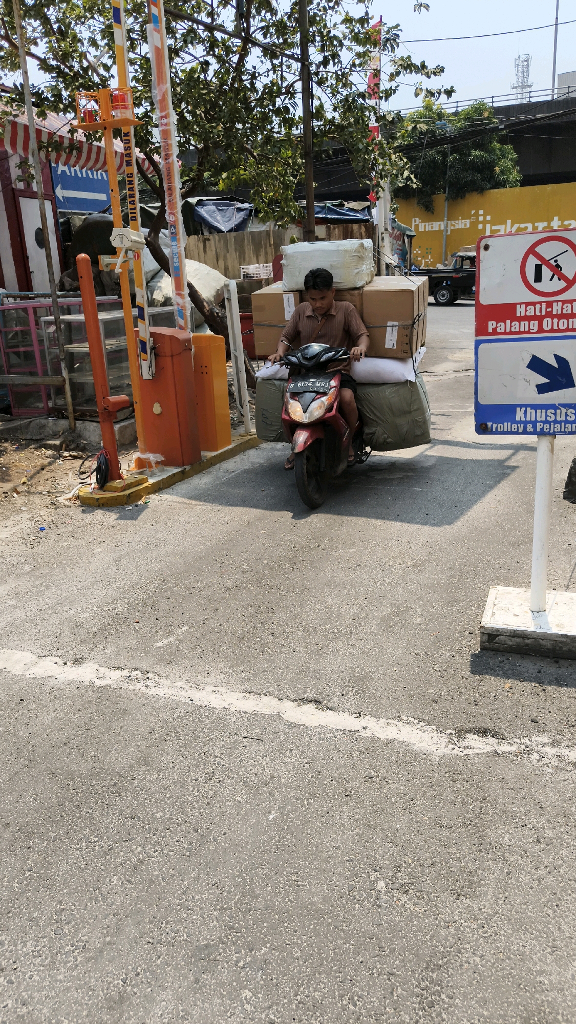 A man is riding a red motorcycle loaded with boxes and large green bags through a toll booth in West Jakarta, Indonesia. He is wearing a brown shirt. The motorcycle is positioned in the center of the image, moving towards the foreground. Behind the motorcycle, there are orange and yellow toll booth structures. To the right of the motorcycle is a white pole with a sign that shows a prohibition symbol and text in Indonesian and English, indicating "Caution - Automatic Barrier" and "For Trolley & Pedestrians". To the left of the motorcycle, there is a blue arrow sign with "LARKIN" written on it. In the background, there are trees, tents, and buildings, including a yellow wall with writing that appears to be "Pinarsia Jakarta". The ground is paved with asphalt. The sky is clear and blue.