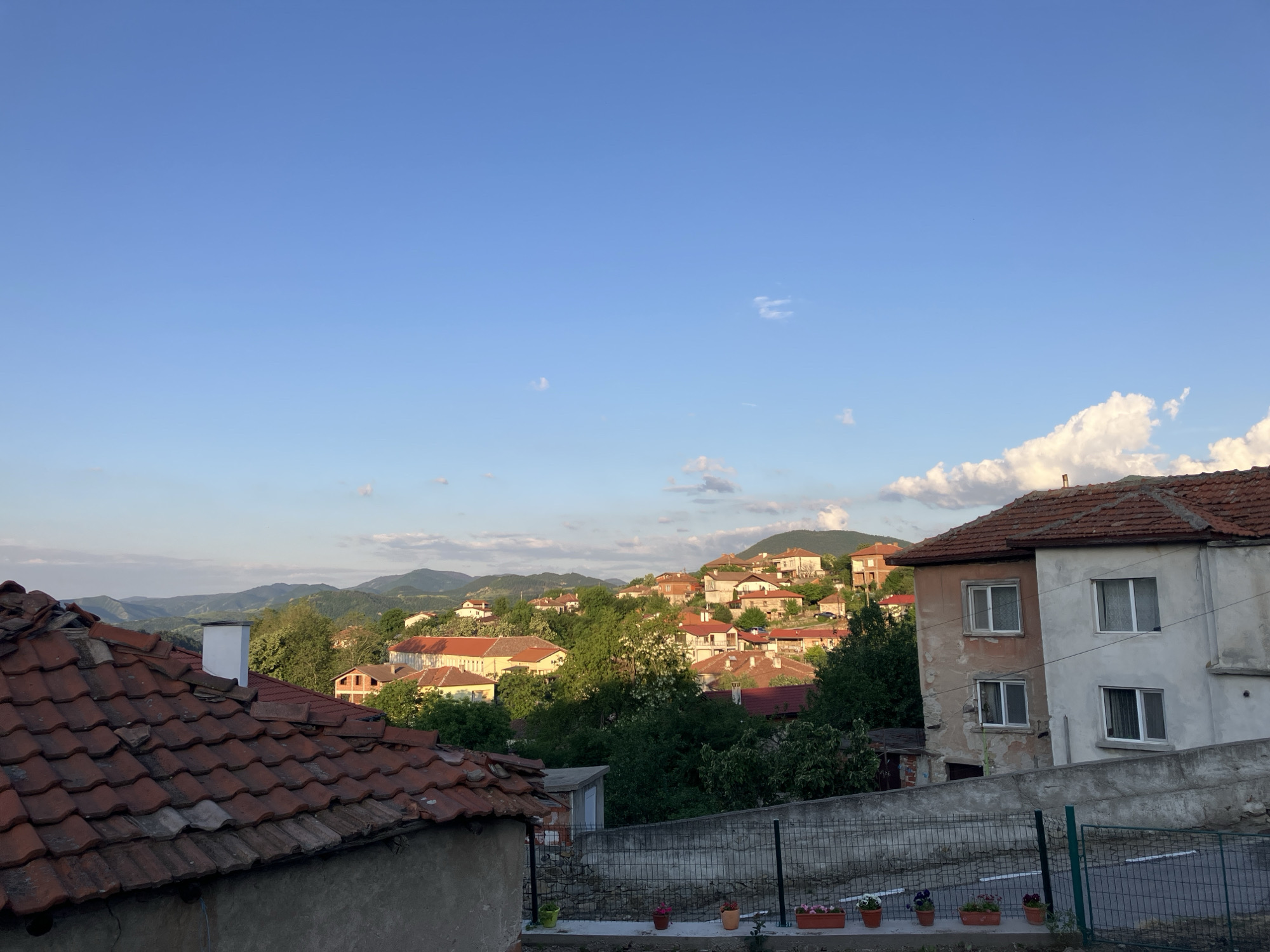 The image shows an outdoor view of Komuniga, Bulgaria. In the foreground are roofs of buildings with red tiles. A two-story building with a red tile roof and peeling white paint is to the right. There is a gray concrete wall with a green metal fence in front of it. Flower pots are lined up along the wall. In the mid-ground, there are houses with red tile roofs situated amongst green trees. In the background, there are mountains against a blue sky with scattered clouds. The overall scene depicts a residential area within a mountainous landscape.