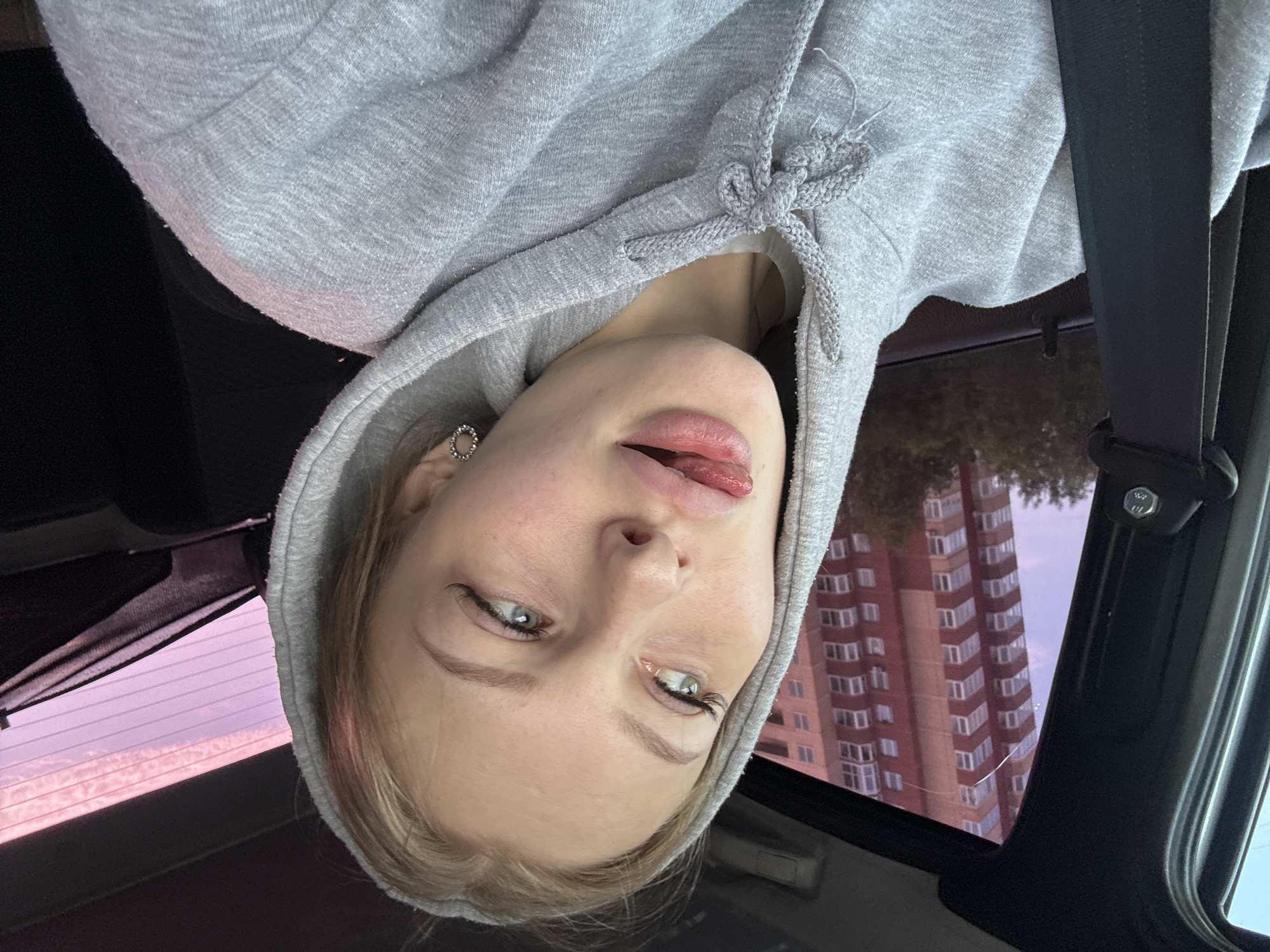 The image is a selfie taken by a young woman, seemingly inside a car. She is wearing a grey hooded sweatshirt, and her hood is pulled up, obscuring her hair to some extent. Her face is the primary subject, with her eyes wide open and looking slightly off-camera. She has her tongue sticking out slightly, with her lips parted, giving a playful or cheeky expression. Her makeup appears to be light, with mascara visible on her eyelashes. She is wearing a circular earring in her visible ear.
The background is blurred and suggests an urban setting. A tall, modern apartment building with many windows is visible through what appears to be a car window. The sky in the background is a light purple hue, suggesting either dawn or dusk. The overall lighting indicates it is daytime. The scene is likely a city in Perm, Russia, as indicated by the location context. The woman's posture and expression suggest a casual and informal moment, possibly while traveling or waiting. There is no visible text in the image.