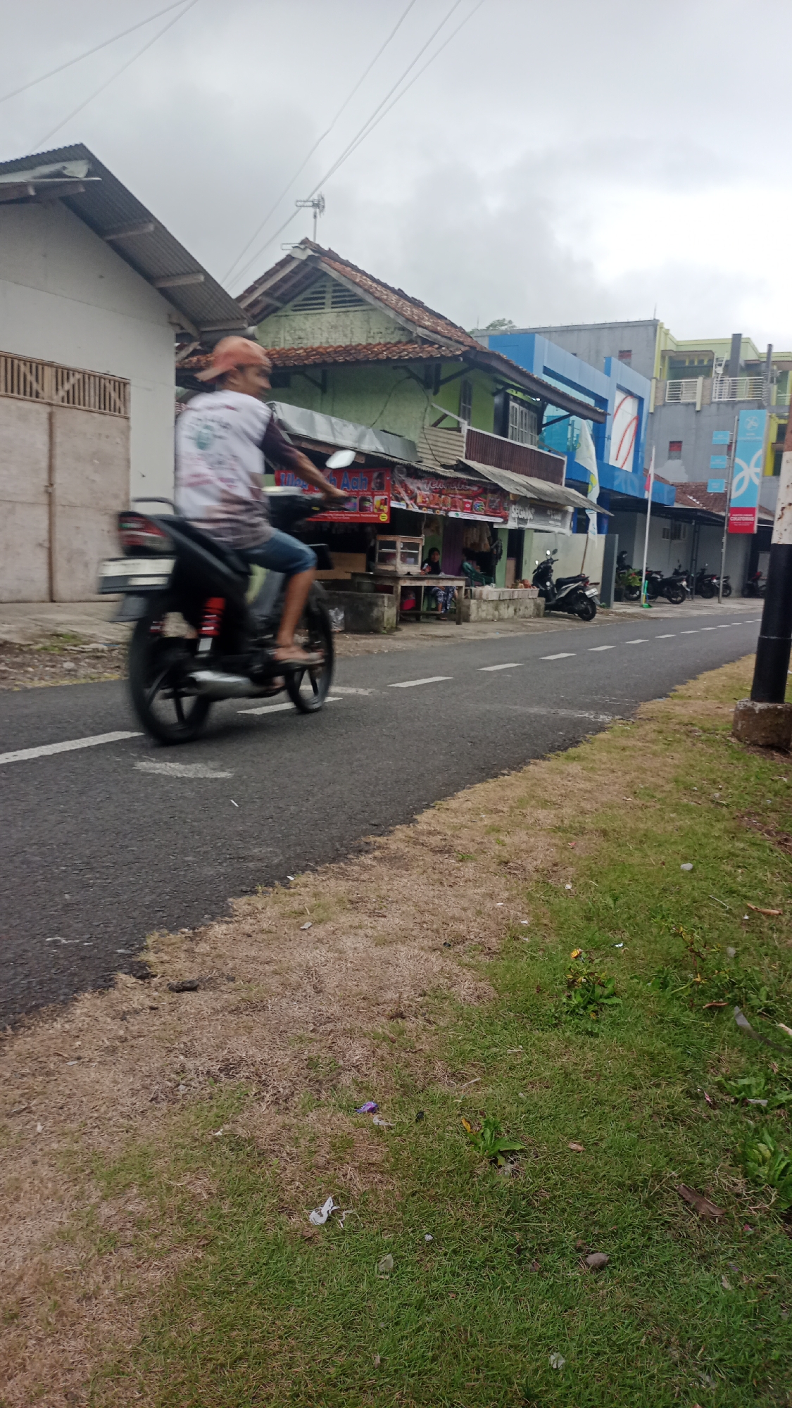A person on a motorcycle is captured in motion on a paved road in Pakemitan, Indonesia. The rider, wearing a cap and a t-shirt, is partially obscured by motion blur. The motorcycle is black with red accents on the suspension. To the right of the road, there is a patch of dry grass followed by a strip of green grass. In the background, there are several buildings. One building on the left has a white facade and a corrugated metal roof. Adjacent to it is a green building with a similar roof, likely a shop. Further down the street, there are modern buildings with blue and gray facades. Several other motorcycles are parked in front of the shops. The sky is overcast.