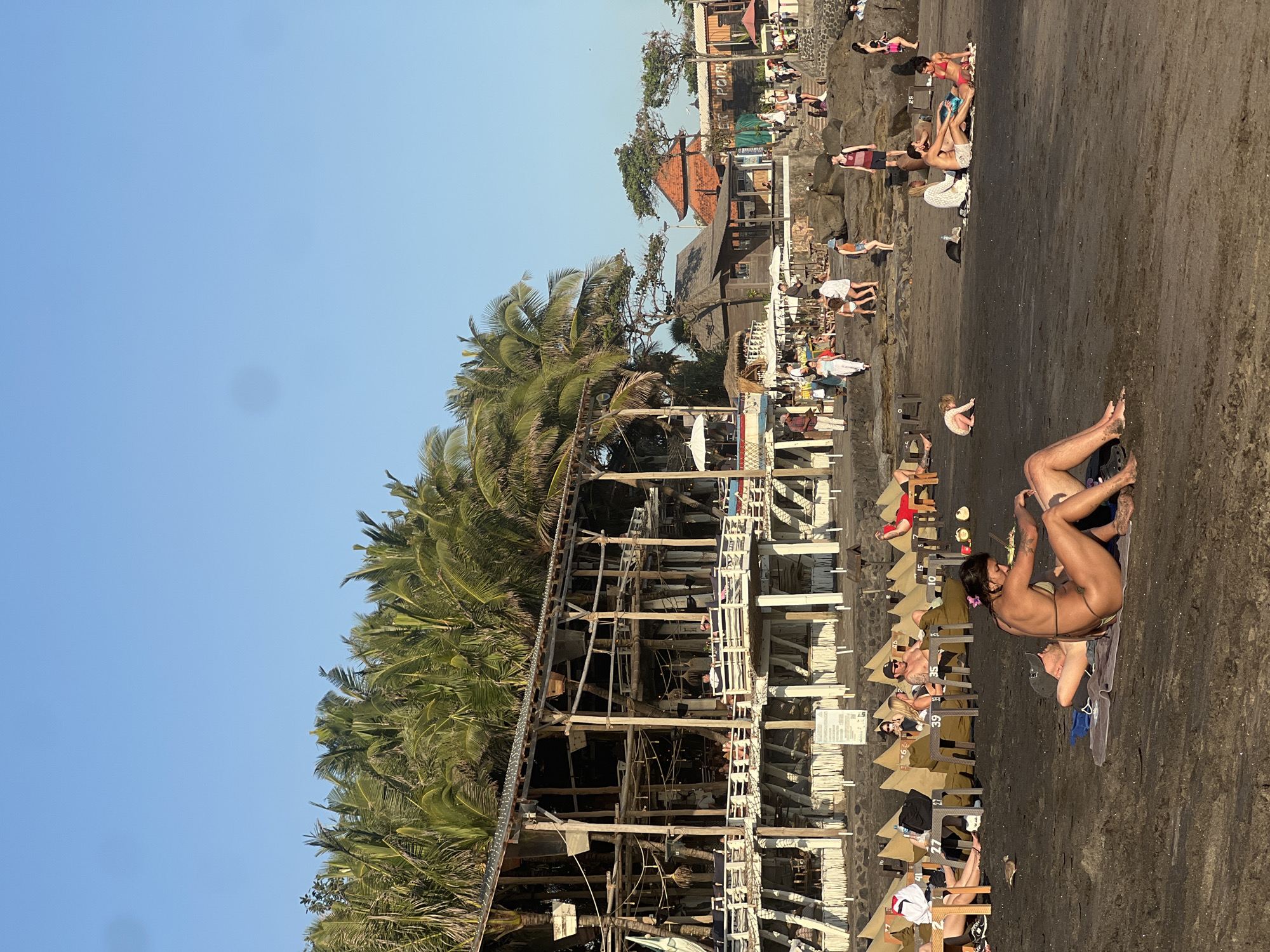 The image depicts a sunny day on a beach in North Kuta, Indonesia. The scene is populated with numerous people enjoying the warm weather. Some individuals are lounging on the dark sand, while others are seated on chairs or relaxing under umbrellas. A few people are visible in the water or near the shoreline.
Dominating the mid-ground of the image is a multi-level wooden structure, likely a beach bar or restaurant, with a thatched roof. It appears to be constructed from natural materials and blends with the tropical environment. Palm trees with lush green fronds are abundant throughout the scene, suggesting a tropical location.
The activity taking place is leisure and recreation at the beach. People are sunbathing, socializing, and enjoying the coastal atmosphere. The time of day appears to be late afternoon or early evening, as indicated by the warm, golden light casting long shadows on the sand. The weather seems to be clear and pleasant, typical for a tropical destination.
In the background, there are more buildings, some with red-tiled roofs, and rocky outcrops. There is also a sign visible on the wooden structure with some text, though it is not clearly legible. The overall emotion conveyed by the scene is one of relaxation and enjoyment, with people making the most of a beautiful day by the sea.