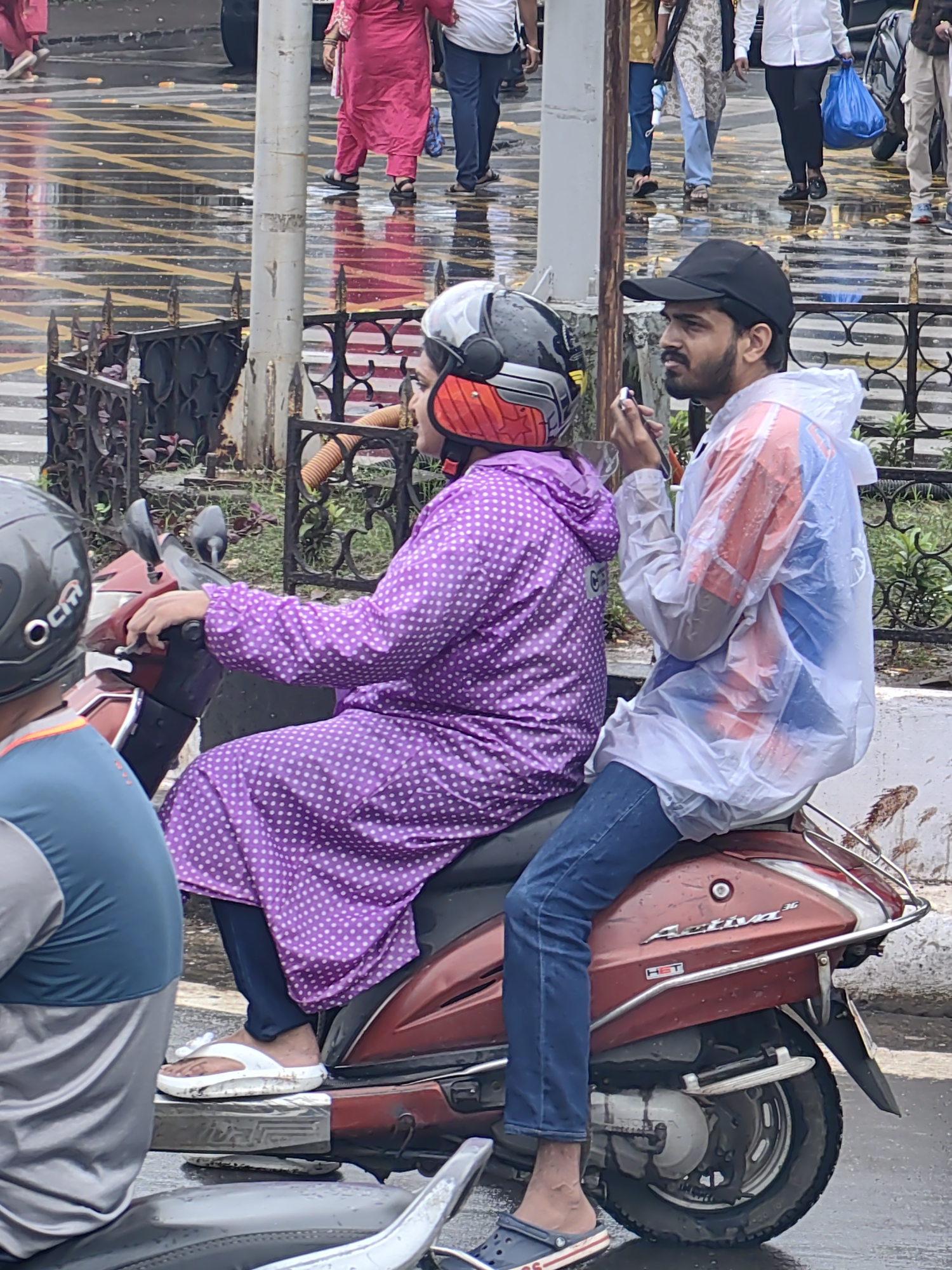 Two people are riding a red scooter on a wet street, likely in Mumbai, India, given the presence of "Activa" branding on the scooter, which is a popular model in India. The scooter has a woman in the front wearing a purple polka-dot raincoat and a silver and red helmet, and a man behind her wearing a white plastic raincoat and a black baseball cap. He appears to be holding a small object in his hand, possibly a cigarette or a phone. The street is wet and reflective, suggesting it is raining or has recently rained. The background shows a sidewalk with some people walking and a metal fence.