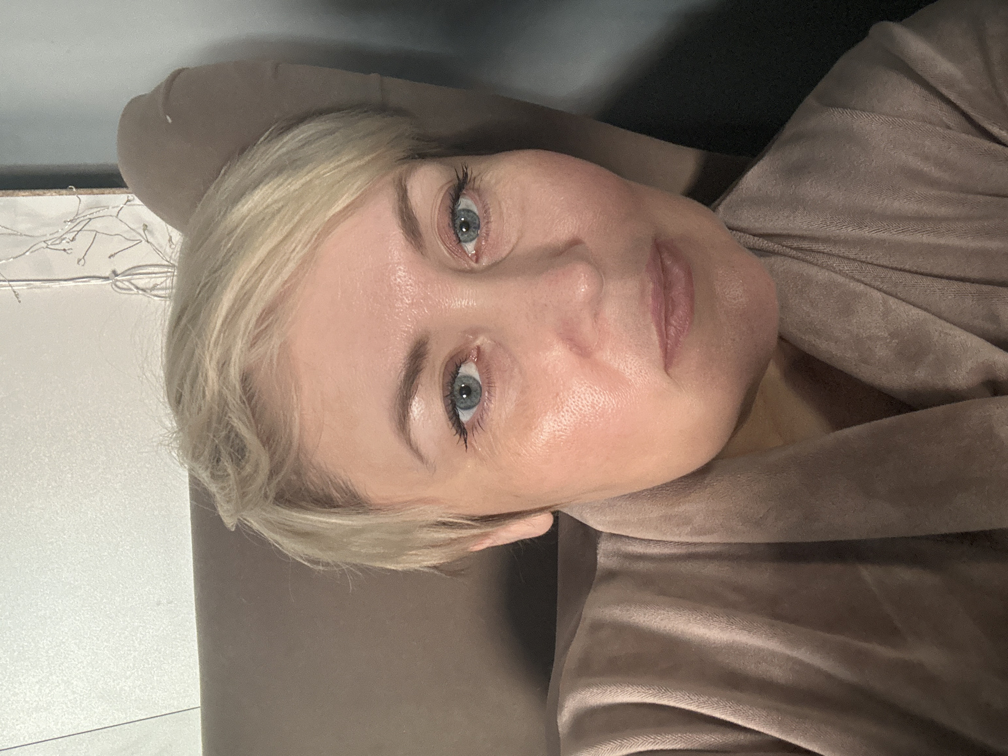 A middle-aged woman with blonde hair and blue eyes is lying on a brown upholstered surface, possibly a couch or chair. She is wearing a brown robe or garment. The background is out of focus, but a white wall with string lights is visible in the upper left. The location is unknown and there is no specific reason to indicate this image was taken in Votkinsk, Russia.