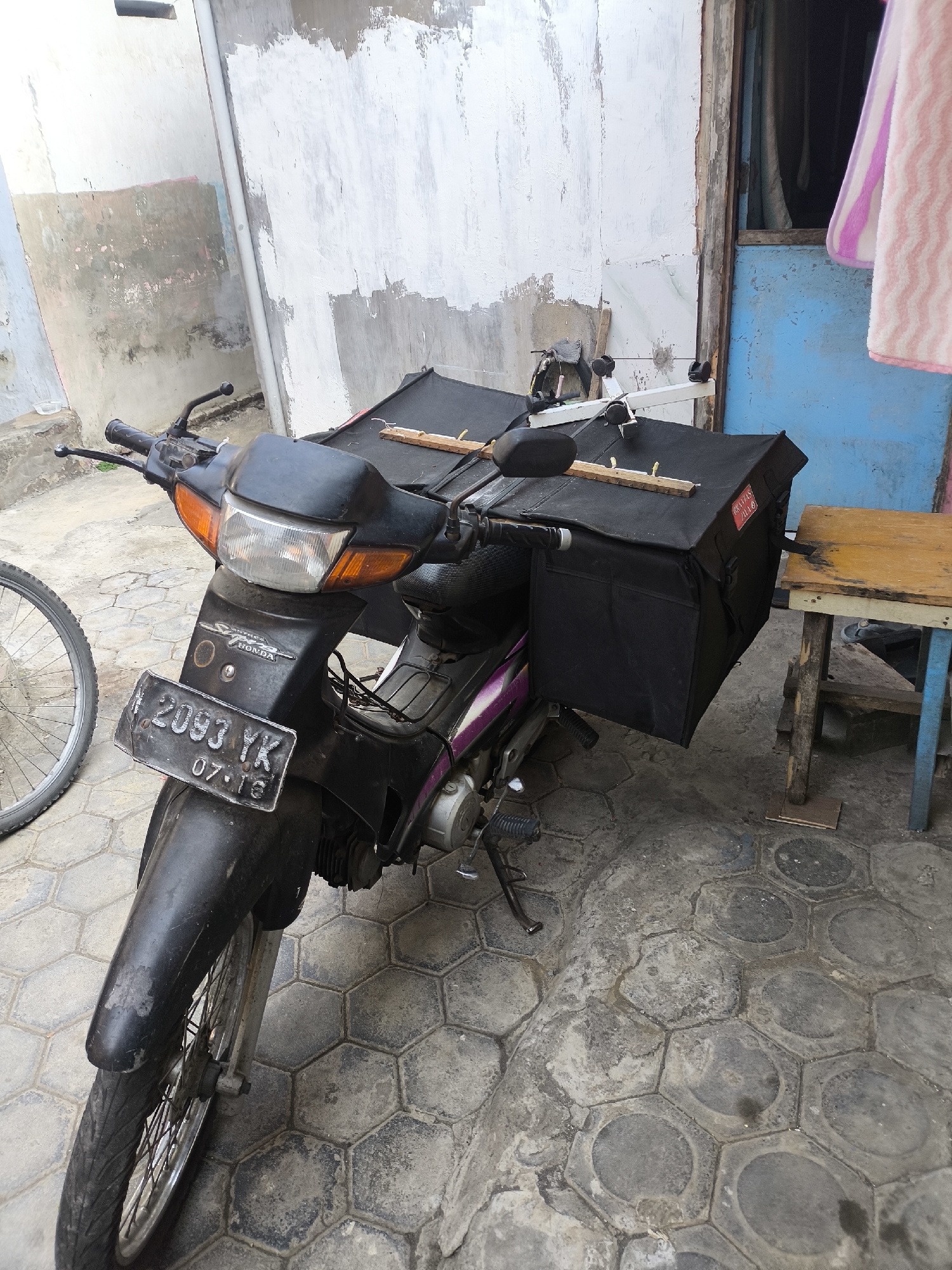 A black motorcycle with large black saddlebags attached to the rear is parked on a hexagonal tiled ground outside a building. The motorcycle has a license plate that reads "2093 YK 07.18". The building's wall is white with patches of grey and pink. To the right of the motorcycle, a small wooden table with blue legs stands near a blue door. A pink and white striped towel hangs on the right side of the building. The scene appears to be an outdoor setting in the city of Citrodiwangsan, Indonesia.