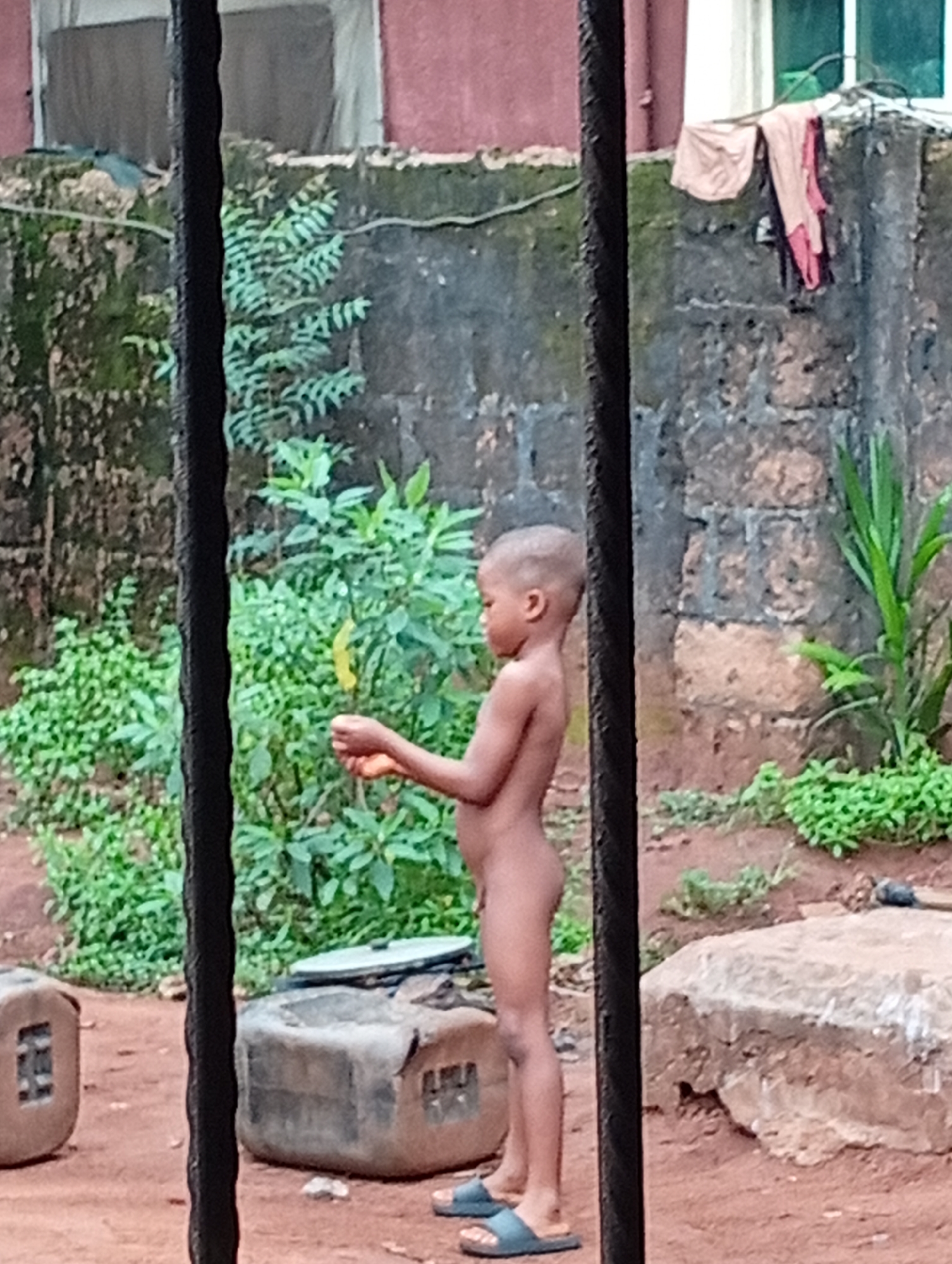 A young child stands outside, nude except for sandals. The child is positioned in a yard with a dirt ground, a stone structure, and various green plants. A building is in the background. The scene is viewed through what appears to be a barred window or gate. Clothing is hanging on the wall.