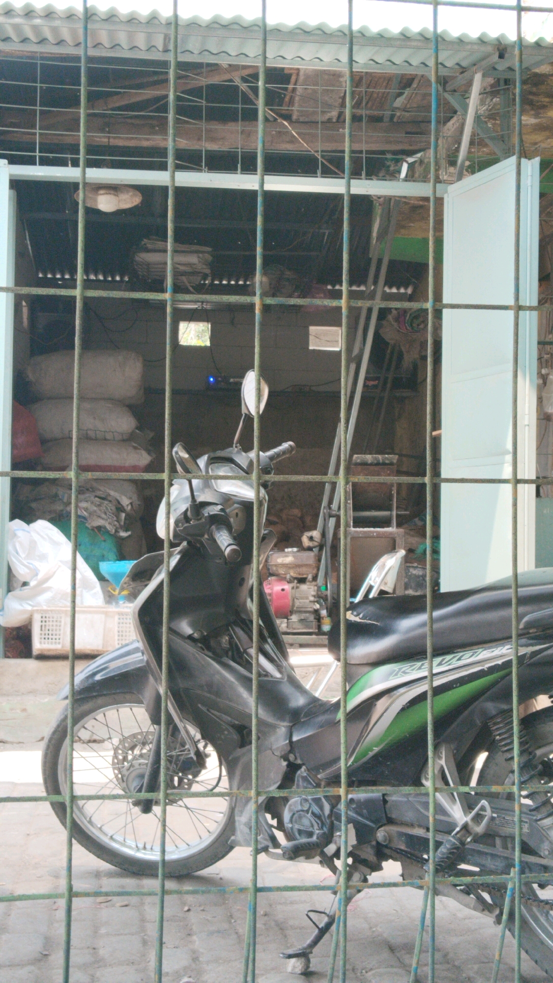 A black motorcycle is parked in front of a storage area with a corrugated metal roof. The motorcycle is viewed through a green metal fence. The storage area appears to be cluttered with bags of what seem to be rice or similar goods, a ladder, and some equipment. A white door is open to the right. The ground is paved. The scene is likely an outdoor area in Losari, Indonesia.