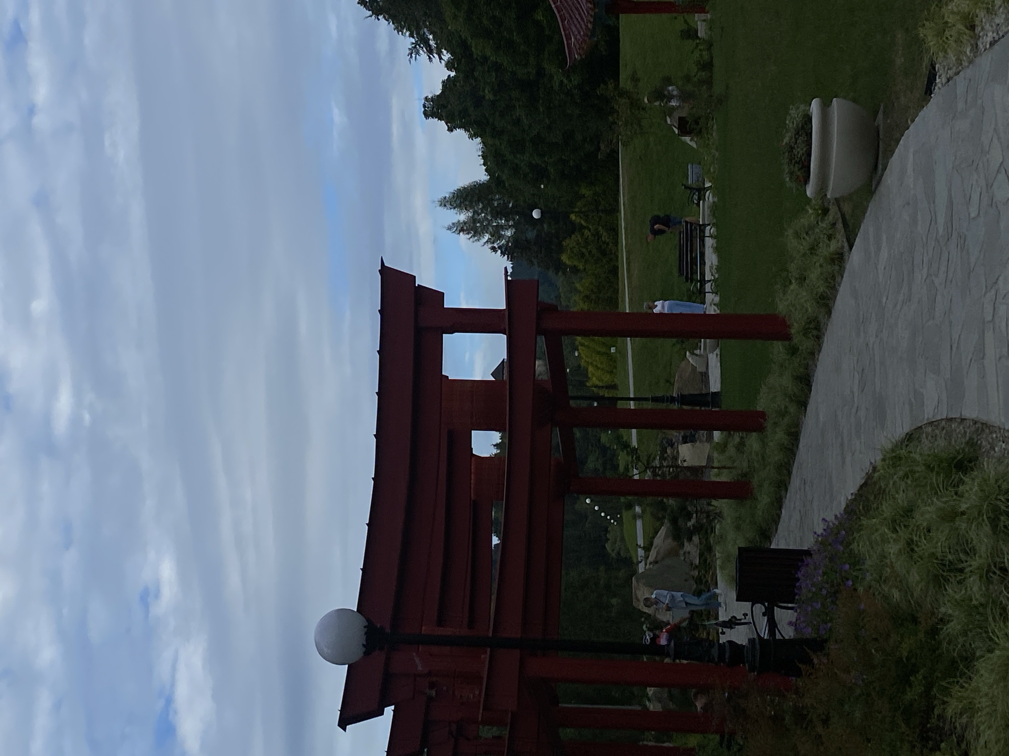 The image shows a Japanese-inspired gate structure painted red, with a path leading around it. There are several people in the background, some standing and some interacting with a bicycle. The scene is set in a park-like environment with trees and greenery, under a cloudy sky. The location is identified as Muszyna, Poland.