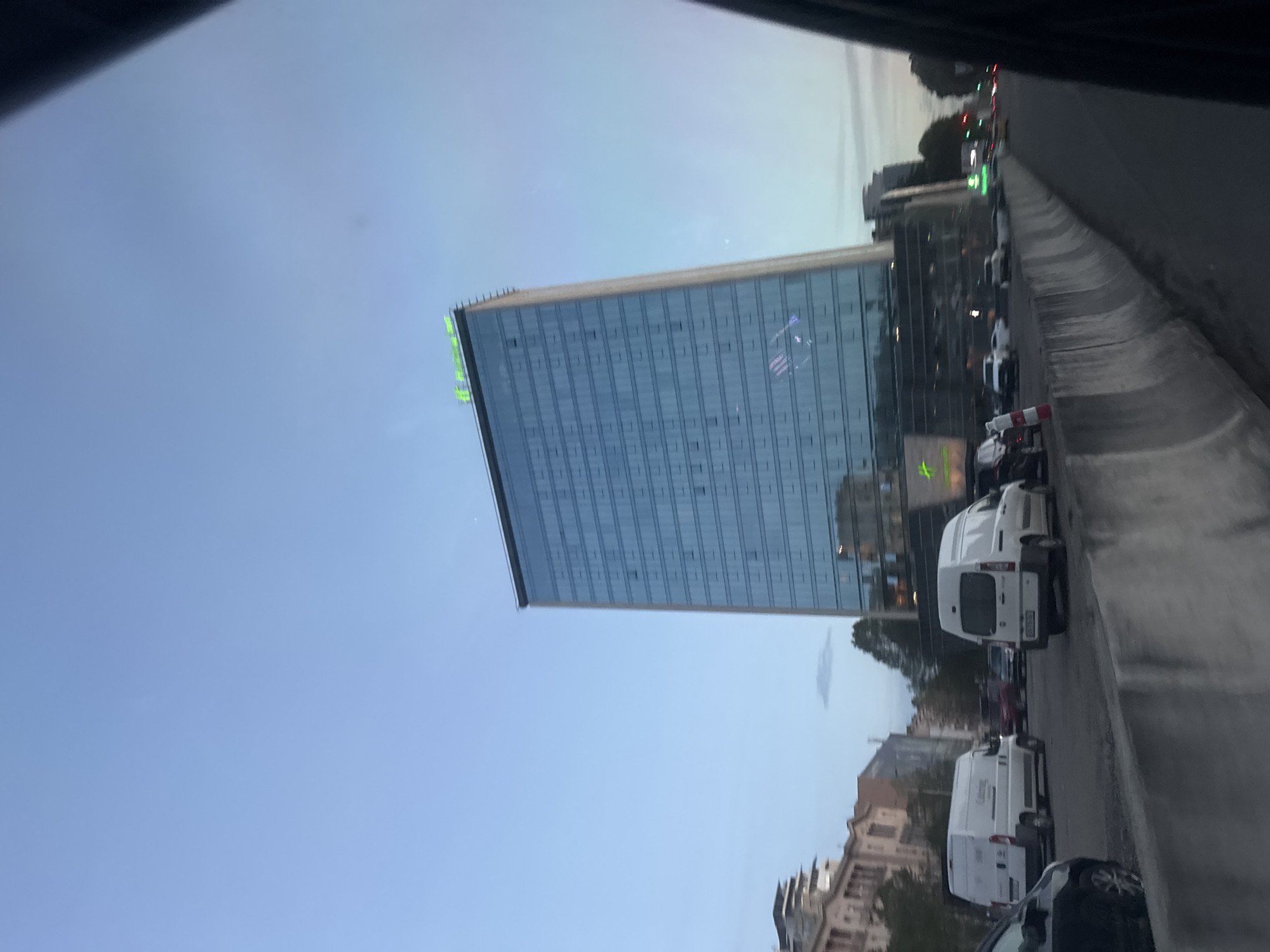 A low-angle view shows a modern glass office building against a clear sky in Tbilisi, Georgia. Cars and vans are parked on a road to the right of the building. Buildings and trees are also visible.