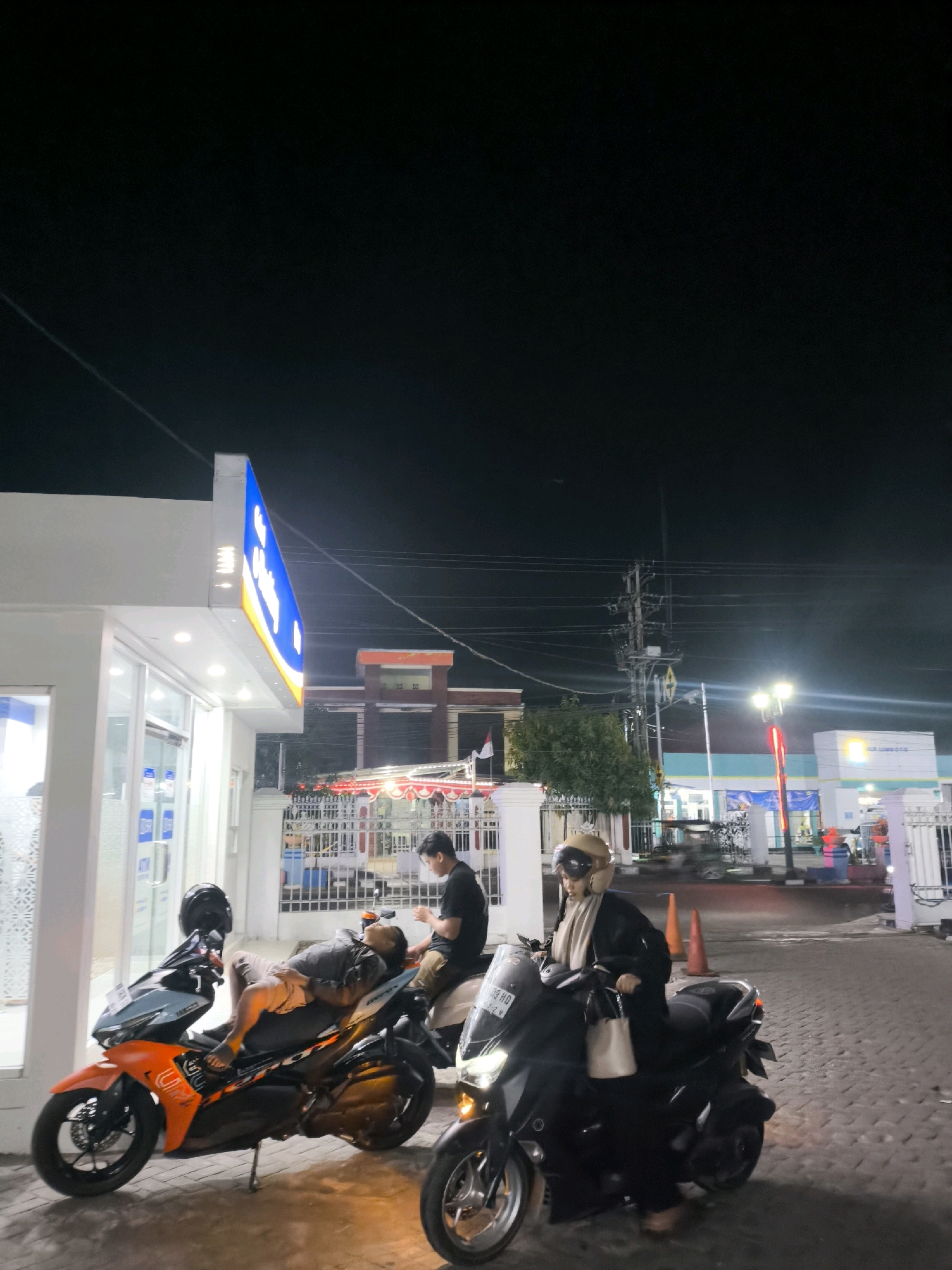 This image captures a night scene at a brightly lit convenience store with a gas station in the background, located in Limboto, Indonesia. A man is lying down on an orange and black motorcycle, appearing to rest or sleep. Another man sits on a different motorcycle behind him, looking towards his phone. A woman wearing a helmet and scarf is seated on a black scooter, holding a light-colored bag. The environment is paved with cobblestones, and various store signs and streetlights illuminate the area. In the distance, there are more buildings and structures, including what appears to be a taller brick building and a smaller, brightly lit structure with a blue sign. Electrical wires and poles are visible against the dark night sky.