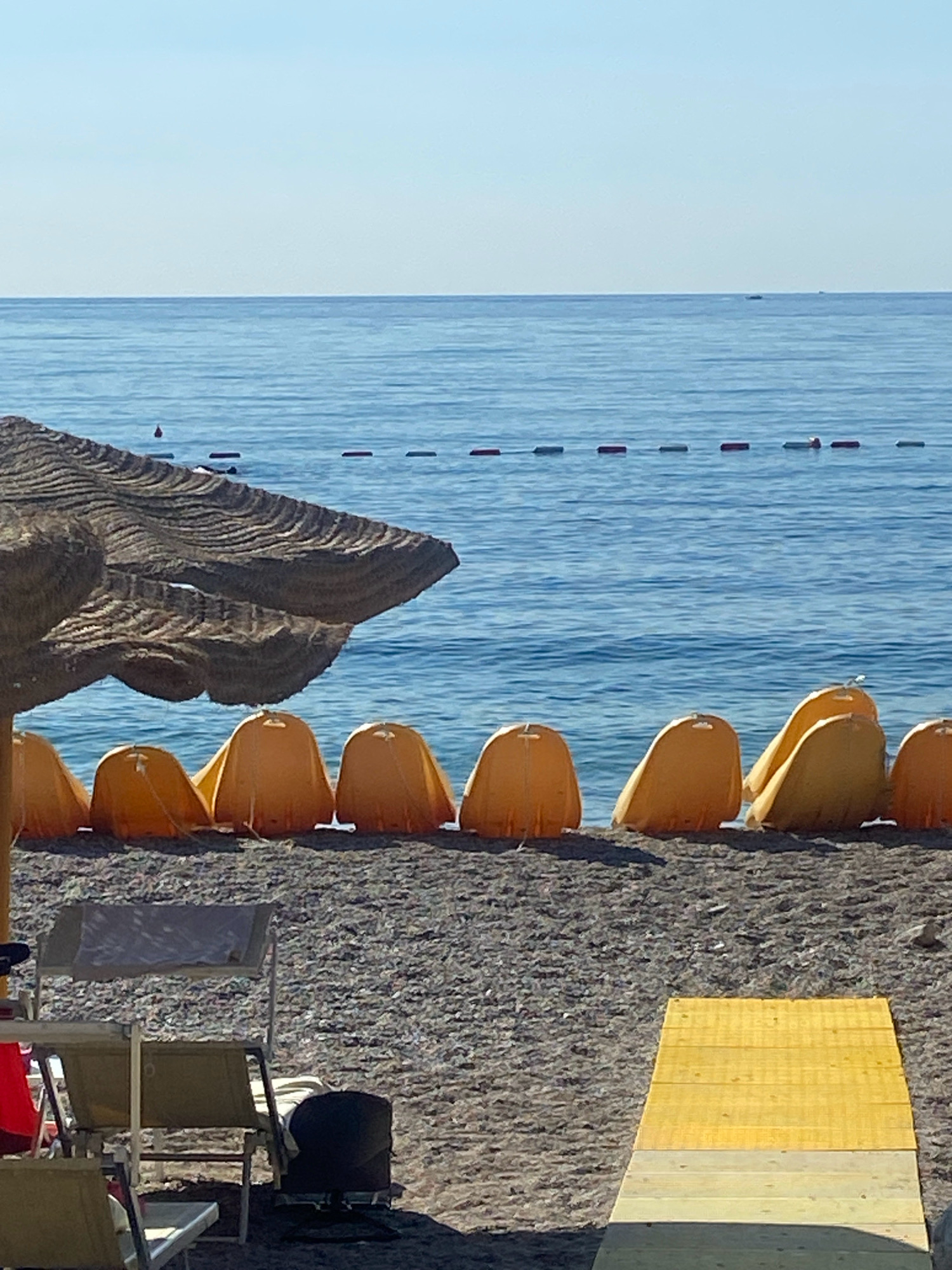 The image depicts a section of a beach in Prijevor, Montenegro. In the foreground, a wooden ramp extends diagonally from the bottom right corner, leading onto the pebbled beach. Several beach chairs, some with fabric seating, are scattered on the sand. A dark object, possibly a trash bin, sits near the chairs.
Further back, a row of bright orange, shell-shaped sunbathers' shields are lined up along the edge of the water. Behind these shields, a large, textured straw umbrella is partially visible on the left side of the frame. The sea stretches out to the horizon, with a calm, blue surface. A line of small buoys or markers is visible on the water. The sky is clear and bright blue. There are no people visible in the image.