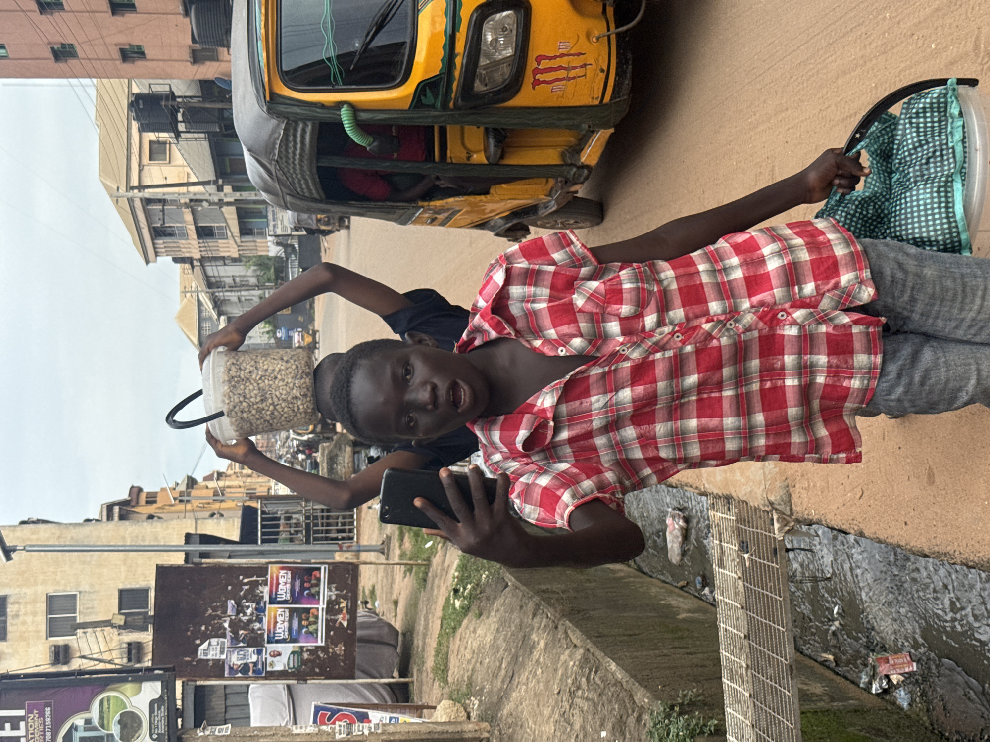 The image captures two young boys in Onitsha, Nigeria. The boy in the foreground is wearing a red and white plaid shirt and gray pants, holding a smartphone and looking towards the camera. His mouth is slightly open, suggesting he might be speaking or reacting to something. Behind him, another boy, wearing a dark shirt, is holding a large clear plastic container filled with small, light-colored objects, possibly nuts or seeds. He is also looking in the direction of the camera.
They are standing on a paved street next to a yellow and black auto-rickshaw. The background shows multi-story buildings, typical of an urban environment in Nigeria. A drainage ditch with flowing water and some debris runs along the side of the street in the foreground. A metal grate covers part of the ditch.
The lighting suggests it is daytime, with an overcast sky. The overall atmosphere appears to be that of everyday street life in a busy Nigerian city.
Visible text includes "WOMEN" and "OPEN MIC" on posters plastered on a bulletin board in the background. There are also some indecipherable words on other signs and posters.