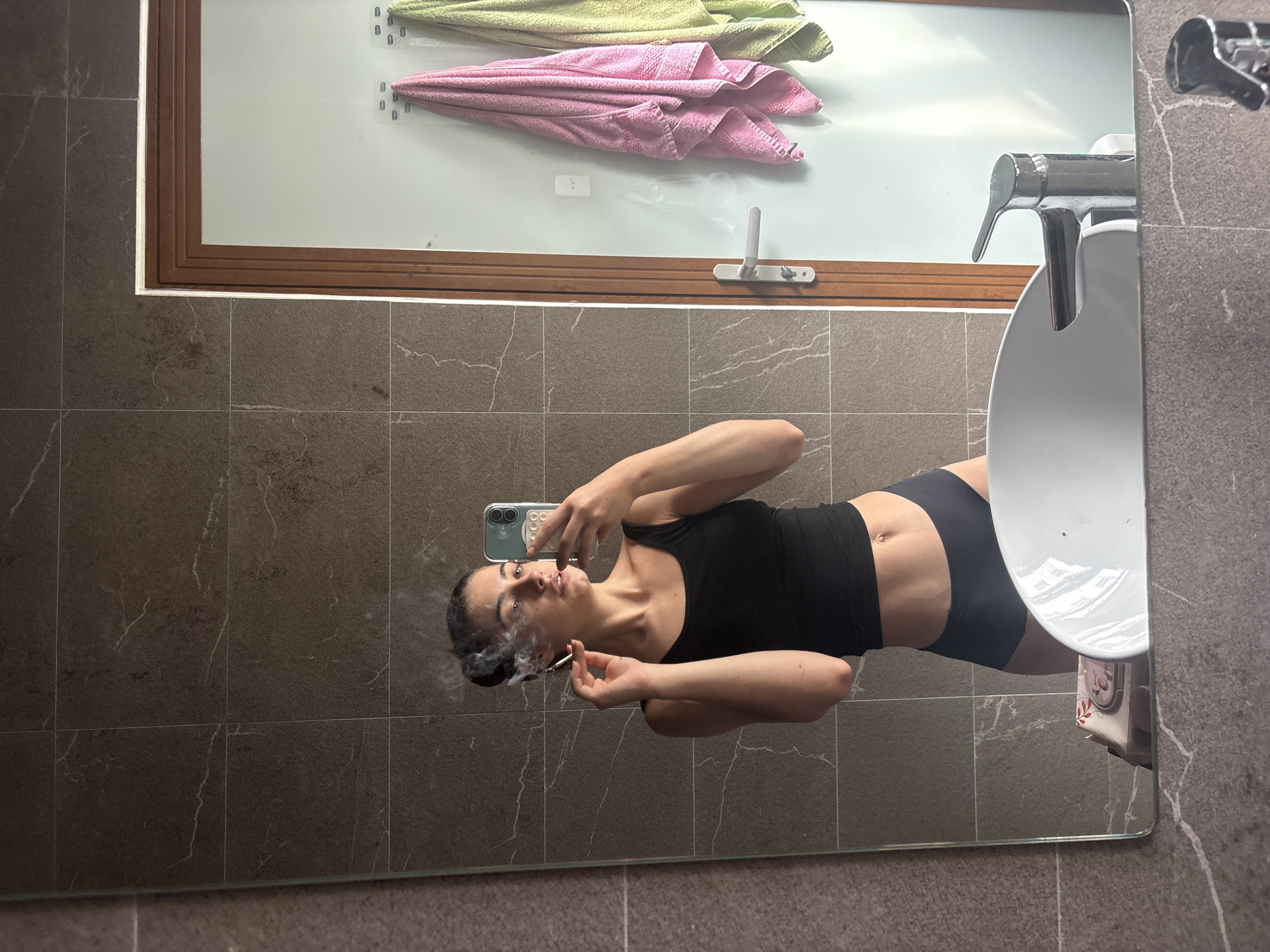 A woman is captured in a mirror selfie within a bathroom setting, appearing horizontally in the frame. She wears a black tank top and dark high-waisted underwear or shorts, exposing her midriff. With her left hand, she holds a light-colored smartphone with a multi-lens camera, pointed towards the mirror to take the photograph. In her right hand, she holds a lit cigarette, from which a distinct plume of smoke is visibly rising around her face as she exhales. Her hair is pulled back, and her gaze is directed towards the phone.
The background, visible in the mirror's reflection, consists of dark grey, stone-patterned tiles with subtle white veining on the walls. Above the main mirror area, a window with a brown wooden frame and a white handle allows natural light into the space. Two towels, one light green and one pink, are draped or hanging near the window. To the right side of the reflection, a white sink and a modern chrome faucet are partially visible. The scene depicts an indoor setting, consistent with a private bathroom, possibly in Đà Nẵng, Vietnam. No discernible text is visible in the image.