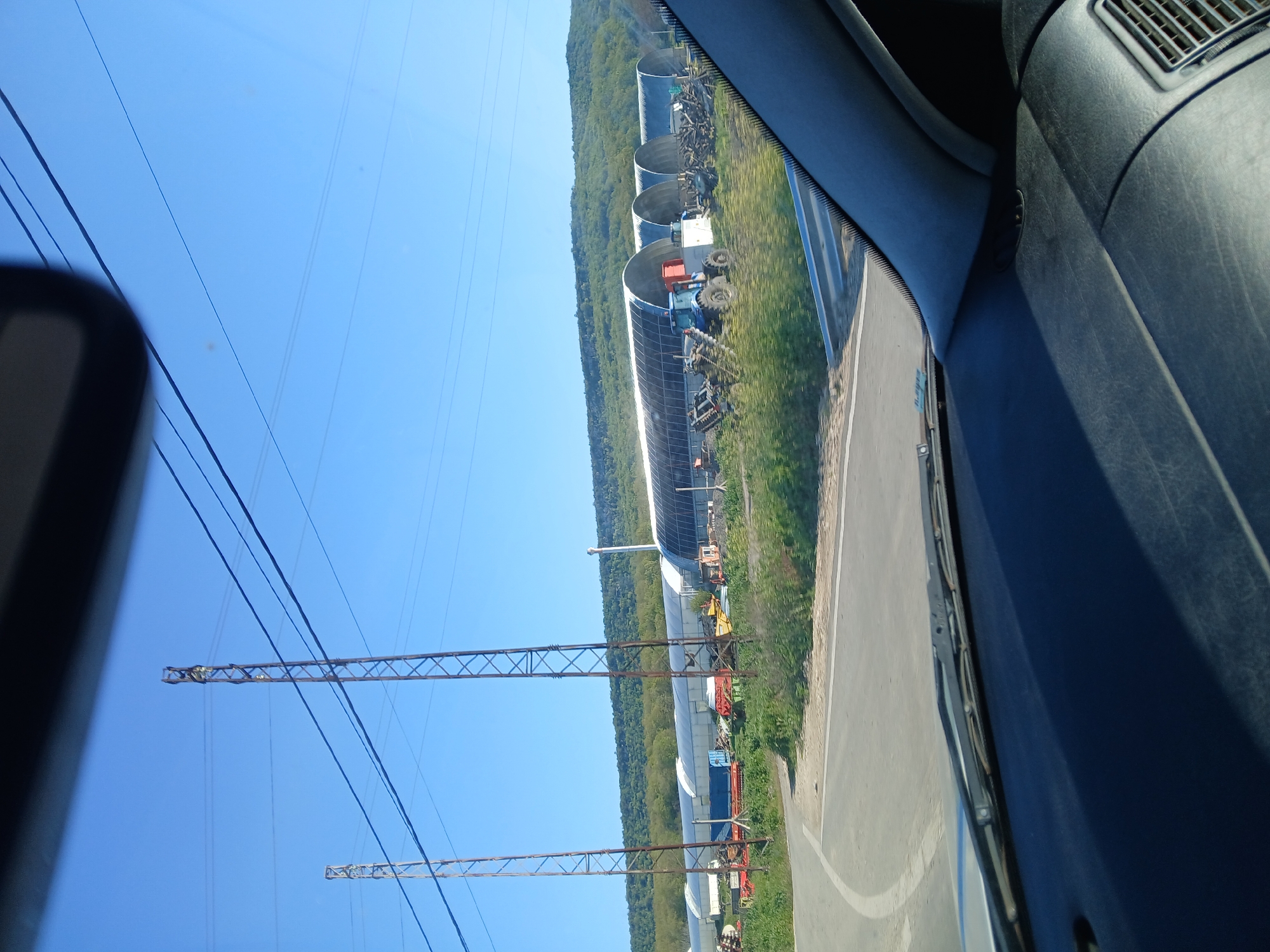 The image shows an outdoor scene from inside a vehicle, possibly a car or truck. The environment consists of a clear blue sky with visible overhead power lines supported by lattice towers. Adjacent to the road is a series of long, arched structures resembling airplane hangars or elongated greenhouses. Various pieces of farm equipment, including tractors, are parked near these structures. The terrain adjacent to the road is grassy with patches of vegetation, backed by a dense line of trees and forest. The location is possibly near Tsarkva, Bulgaria, based on the presence of agricultural structures and terrain characteristic of that region.
