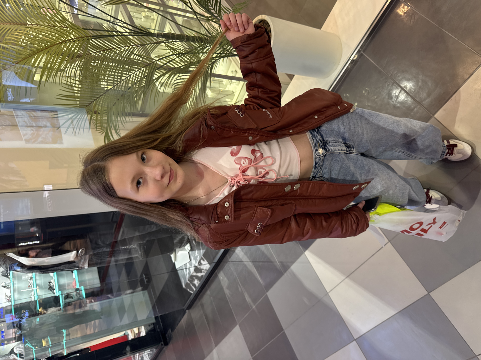 A young girl stands on a checkered tiled floor in what appears to be a shopping mall. She is wearing a brown jacket over a white top with a pink design, blue jeans, and sneakers. She is holding a strand of her long, light brown hair in her right hand. In her left hand, she is holding a white shopping bag with red lettering and a hint of a bright yellow item inside. Behind her, there is a display case with various items on shelves and a large potted plant. The image suggests an indoor environment with modern flooring and potentially commercial establishments nearby. While the image provides visual details, determining the specific location in Ol'ginskaya, Russia, is not possible without additional contextual information.