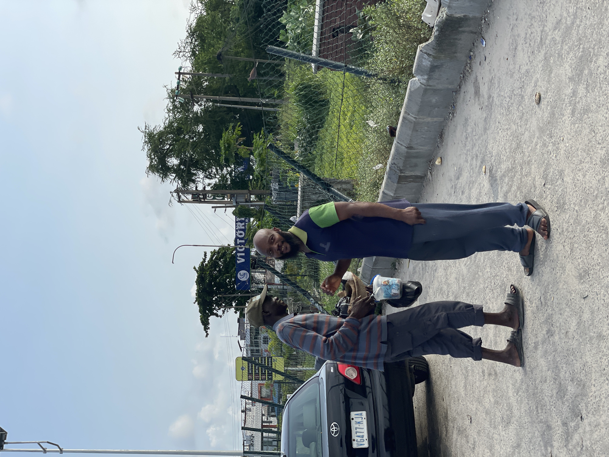 Two men stand on a paved area with a black Toyota Corolla car on the left. The man on the left wears a striped long-sleeved shirt, dark pants, and a cap. He holds a woven basket and a white plastic container with items inside. The man on the right wears a dark blue polo shirt with a green collar and dark pants. He has a beard and is smiling. Behind them, a chain-link fence encloses an area with dense green vegetation and trees. A sign reading "VICTORY" is visible in the background, along with utility poles and wires. The scene appears to be a street in Lekki, Nigeria, under a bright, cloudy sky.