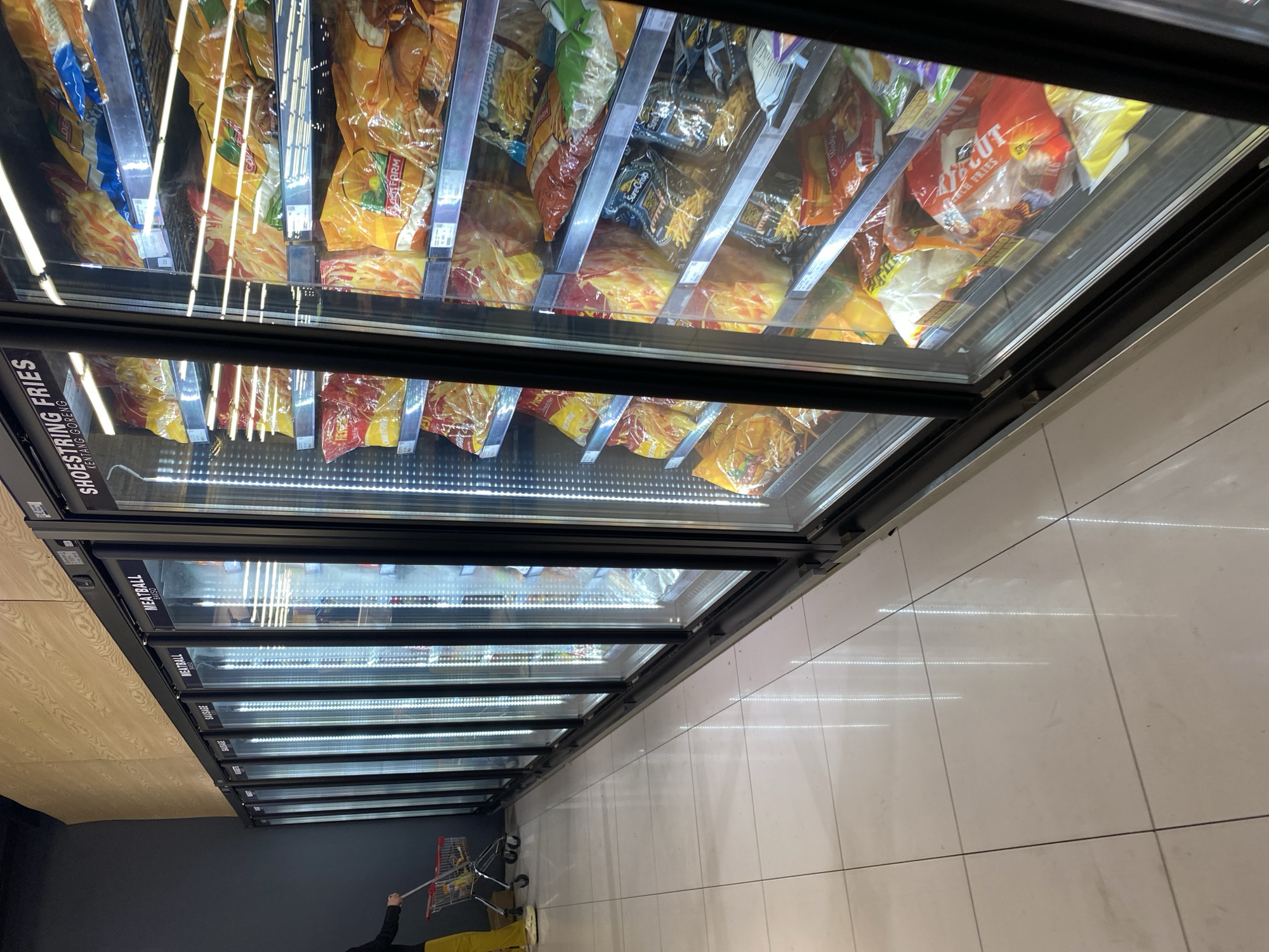 The image shows a row of open-door refrigerators in a grocery store, likely in Banda Aceh, Indonesia. The refrigerators are stocked with various frozen food products, primarily bags of french fries and meatballs. The lighting inside the refrigerators is bright, illuminating the products on the shelves. A person in yellow pants and a dark shirt is visible on the left side of the image, pushing a shopping cart. The floor of the store is tiled with light-colored squares, reflecting the light from the refrigerators. The store appears clean and well-organized.