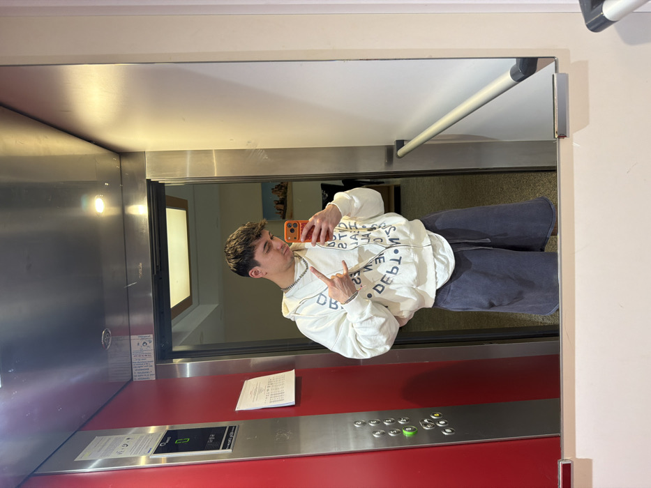 A young adult male is captured in a mirror selfie inside an elevator, located in Sofia, Bulgaria. The male subject, with short brown hair, faces the mirror, winking with his left eye while holding an orange-cased smartphone in his right hand. His left hand forms a "shaka" gesture. He is dressed in a cream-colored, zippered hooded sweatshirt featuring inverted dark text, a white t-shirt, dark grey knee-length shorts, a silver chain necklace, and a silver bracelet on his left wrist.
The mirror's reflection reveals the elevator's interior and a portion of the area outside its open doors. The left side of the reflection shows a red elevator control panel with a green digital display indicating "0" and a vertical column of silver buttons, one of which is illuminated green. White paper notices are affixed to the red panel. Through the open metallic silver elevator doors, a hallway or lobby area is visible, featuring a light-colored ceiling with a square light fixture, a wall-mounted landscape painting depicting rock formations, and a dark piece of furniture, possibly a sofa. The floor visible through the open doors is a dark grey carpet. The right side of the reflection shows a plain white wall with a horizontally mounted silver handrail. The elevator's ceiling is metallic with integrated lighting.
