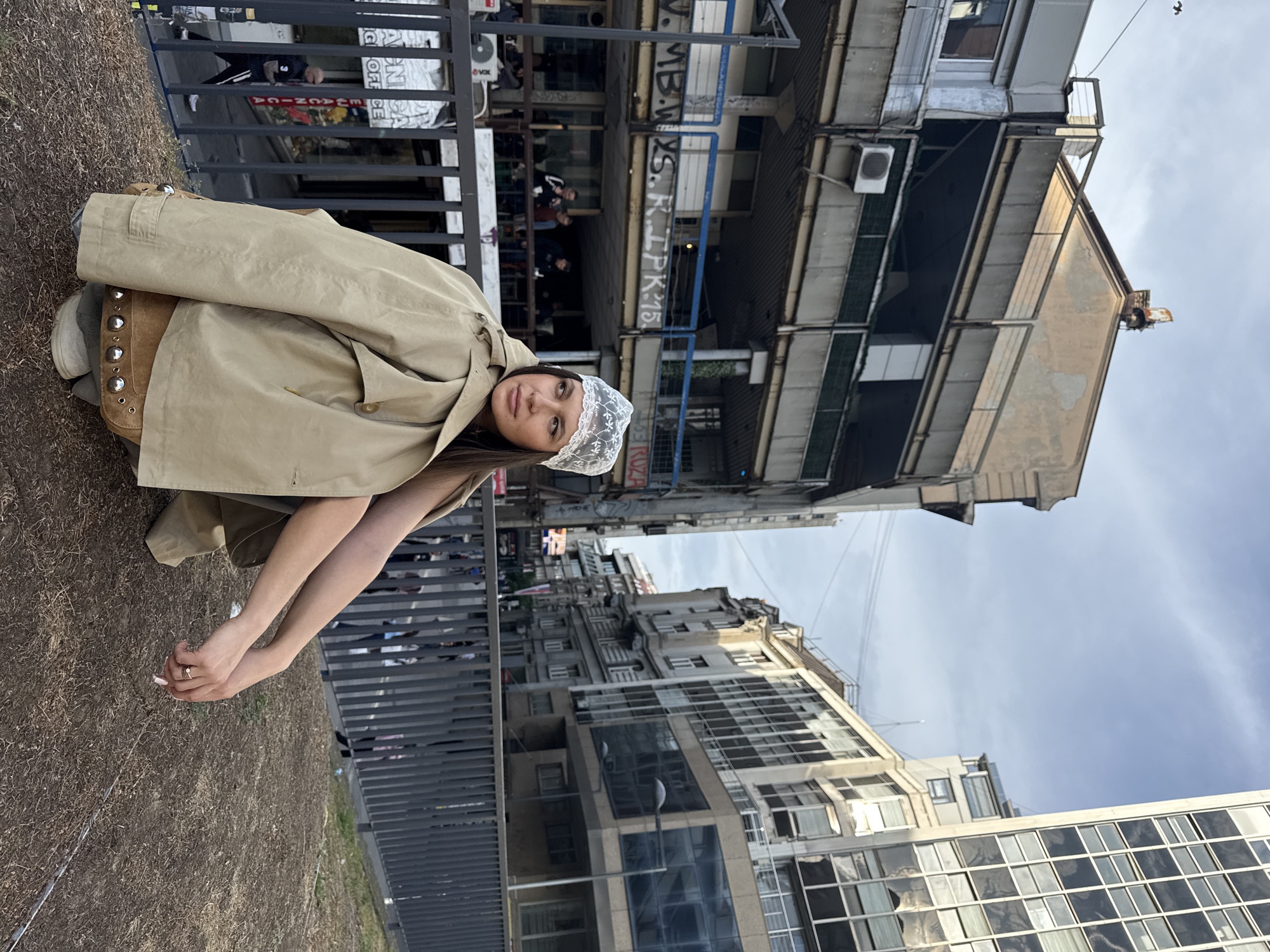 The image captures a young woman in a beige trench coat and a delicate lace head covering sitting on the ground near a metal railing. The woman has dark hair and appears to be posing for the photograph. Behind her, an urban landscape rises vertically, consisting of a variety of buildings with different architectural styles. One building is particularly noticeable with its weathered, multi-storied facade and graffiti on the side. People are visible in the background, walking behind the railing near the building. The sky is overcast with a slight cloud cover. Based on visual cues such as architectural styles, graffiti, and the text on the walls, the location appears to be in Belgrade, Serbia.