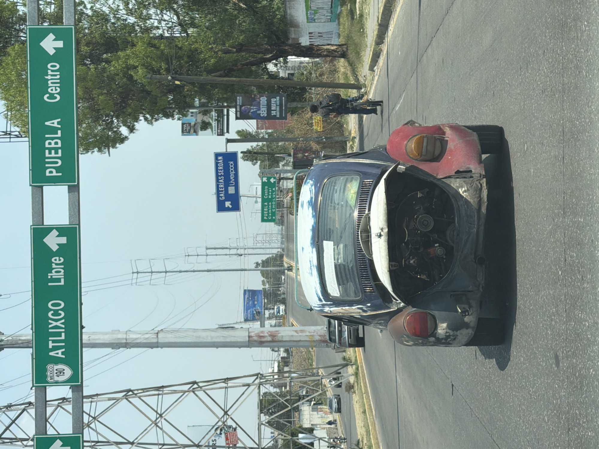 The image shows a road scene in San Bernardino Tlaxcalancingo, Mexico. A damaged blue Volkswagen Beetle with its hood open is stopped on the side of the road. The rear right portion of the vehicle is red and appears to have some damage. Road signs are visible, indicating directions to Atlixco, Puebla Centro, and Galerías Serdán/Liverpool. Power lines stretch across the sky. Further down the road, a man is riding a motorcycle. Banners or signs with text and images are visible along the side of the road.