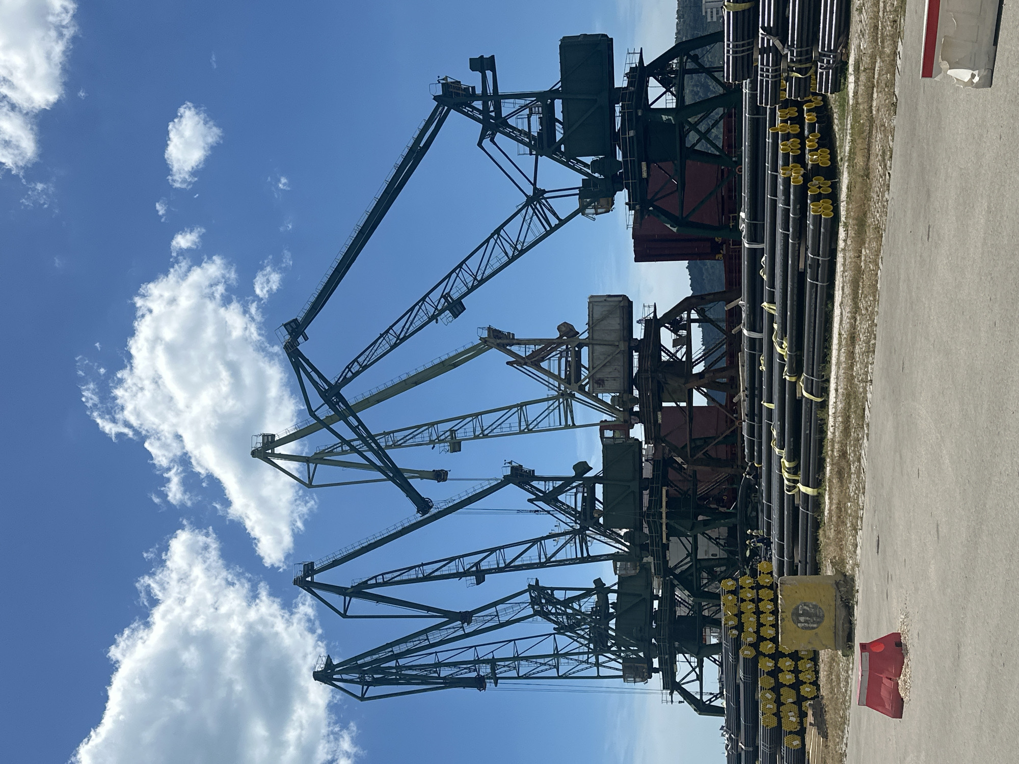 A low-angle, medium shot captures a bright, sunny day in a port with multiple large, dark green industrial cranes standing tall against a vibrant blue sky dotted with fluffy white clouds. In the foreground, neatly stacked bundles of dark metal pipes, many with bright yellow markings, are arranged on the ground. A red concrete barrier and a yellow concrete block with the number "112" stenciled in black are also visible. The cranes, industrial in design with intricate metal frameworks, are positioned behind the pipes. Some cranes have visible operator cabs. In the background, hints of other industrial structures and possibly a hillside with trees can be seen. The overall scene depicts an active industrial port.