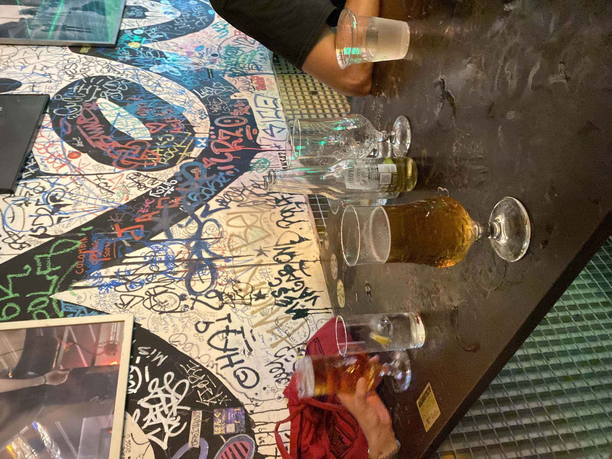 The image depicts a dimly lit interior scene, likely a bar or restaurant, with a prominent graffiti-covered wall in the background. The wall is a canvas of colorful and intricate graffiti art, featuring a mix of styles, text, and abstract designs. In the foreground, a dark, reflective surface, presumably a bar counter, is visible. Several glasses are placed on the counter, containing beverages, including a glass of amber-colored liquid (likely beer) in the foreground. A red bag is partially visible on the left side of the counter. To the right, a person's arm and shoulder are visible, suggesting their presence at the bar. The overall atmosphere suggests a casual, urban setting. The graffiti art on the wall includes various tags and phrases. One discernible text on the wall appears to be "COLOMBIA COMING SOON". There is no specific indication within the image that places the location in Barcelona, Spain.
